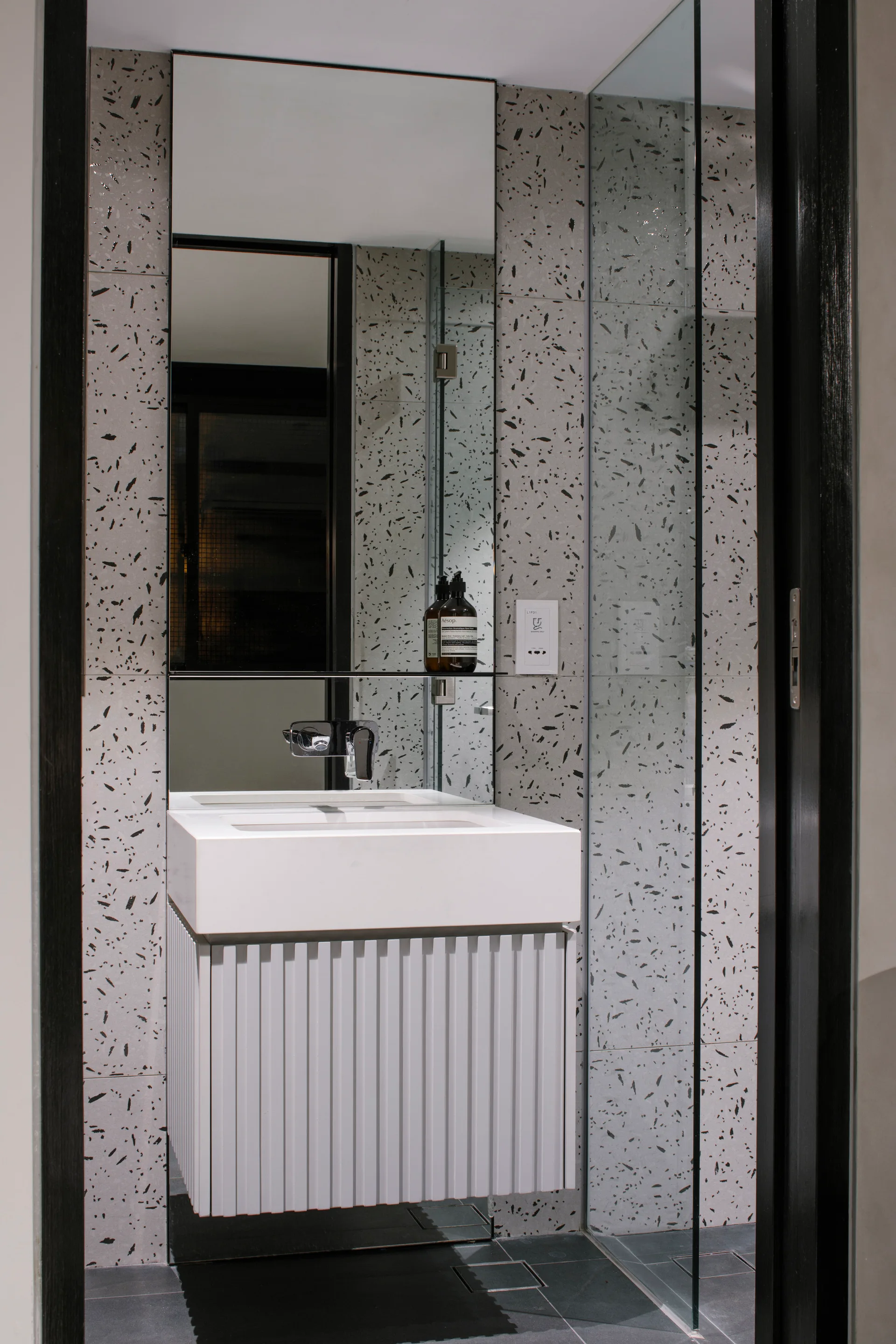 A minimalist bathroom features a white fluted vanity with a chrome wall-mounted faucet set against grey speckled terrazzo-style wall tiles.