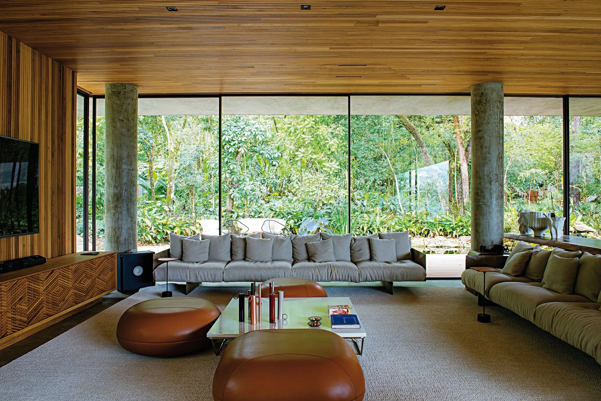 A modern living room features low-profile sofas, leather ottomans, and raw concrete pillars before a floor-to-ceiling glass wall with forest views.
