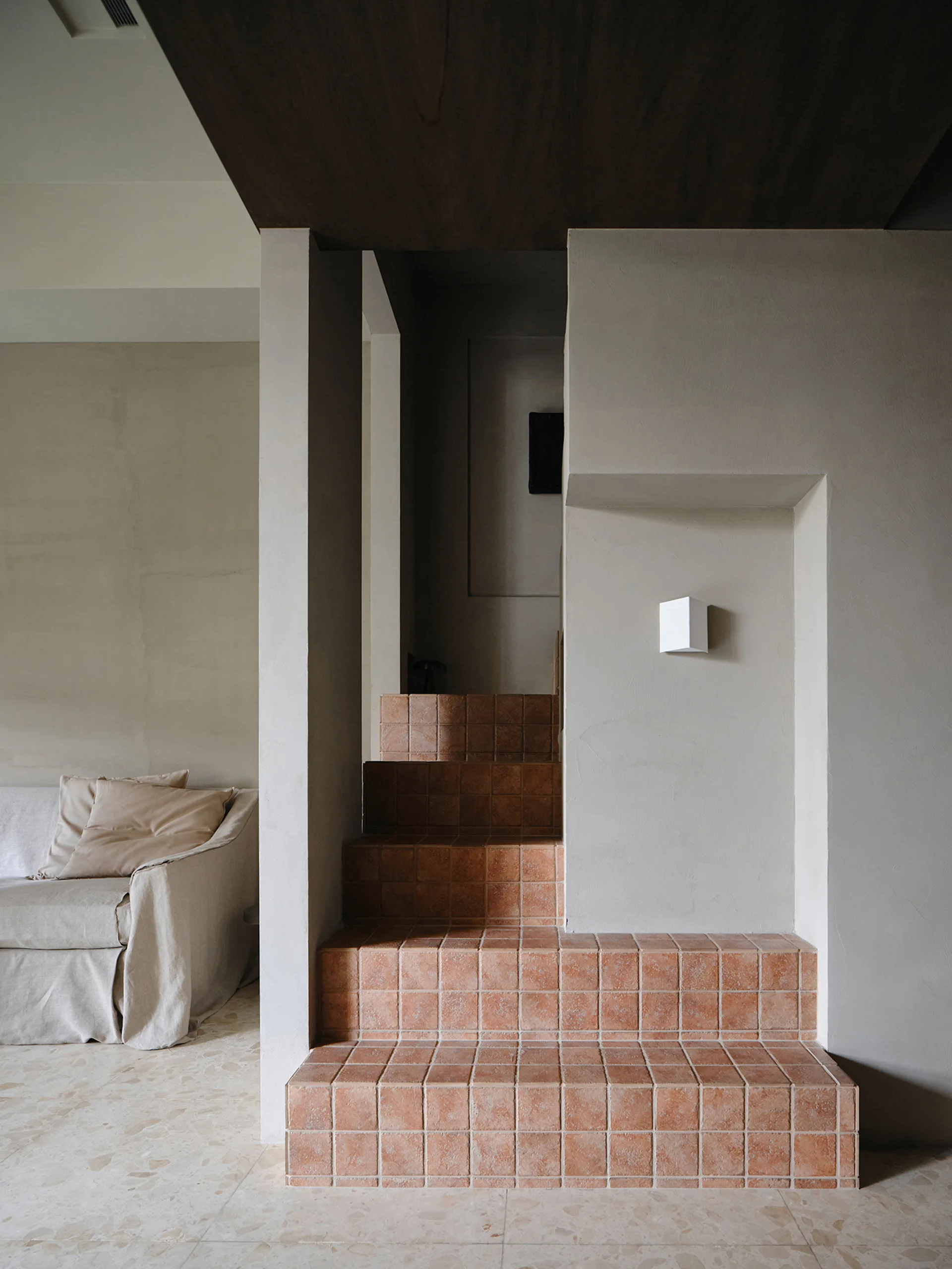 A minimalist interior featuring a staircase covered in square terracotta tiles next to a slipcovered beige sofa and matte plaster walls.