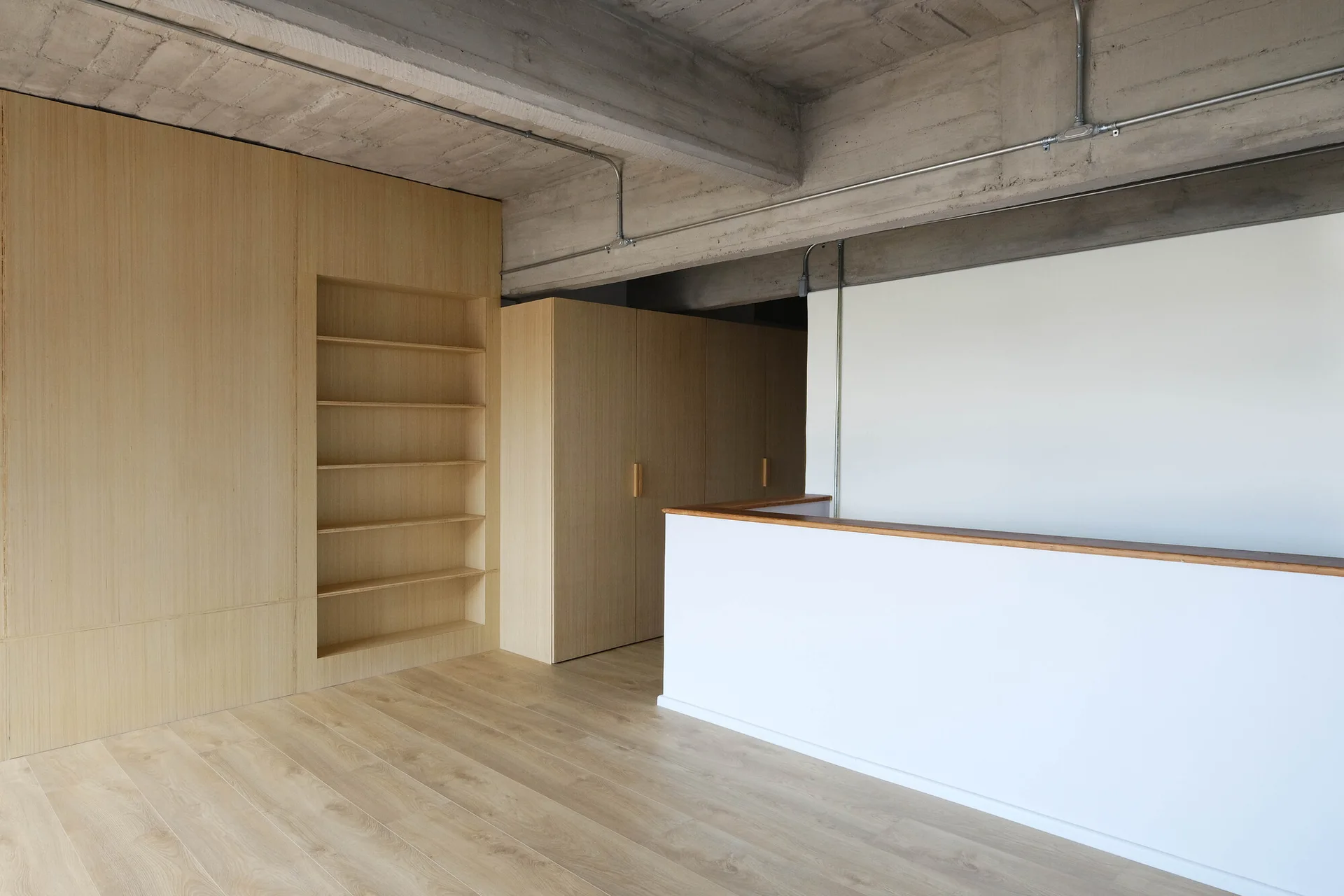 A minimalist room features an exposed concrete ceiling with metal conduits, light wood paneled built-in shelving, and a white partition wall.
