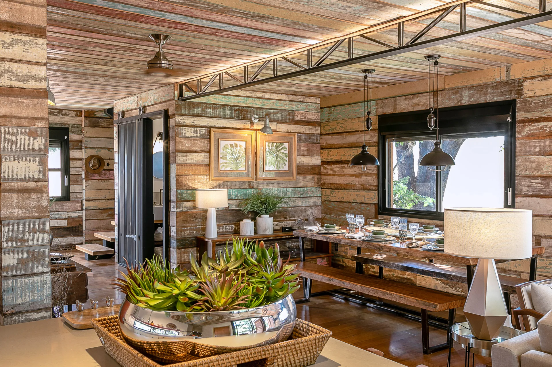A rustic dining room with weathered wood paneled walls, a matching wood ceiling, a dining table with benches, and industrial pendant lights.
