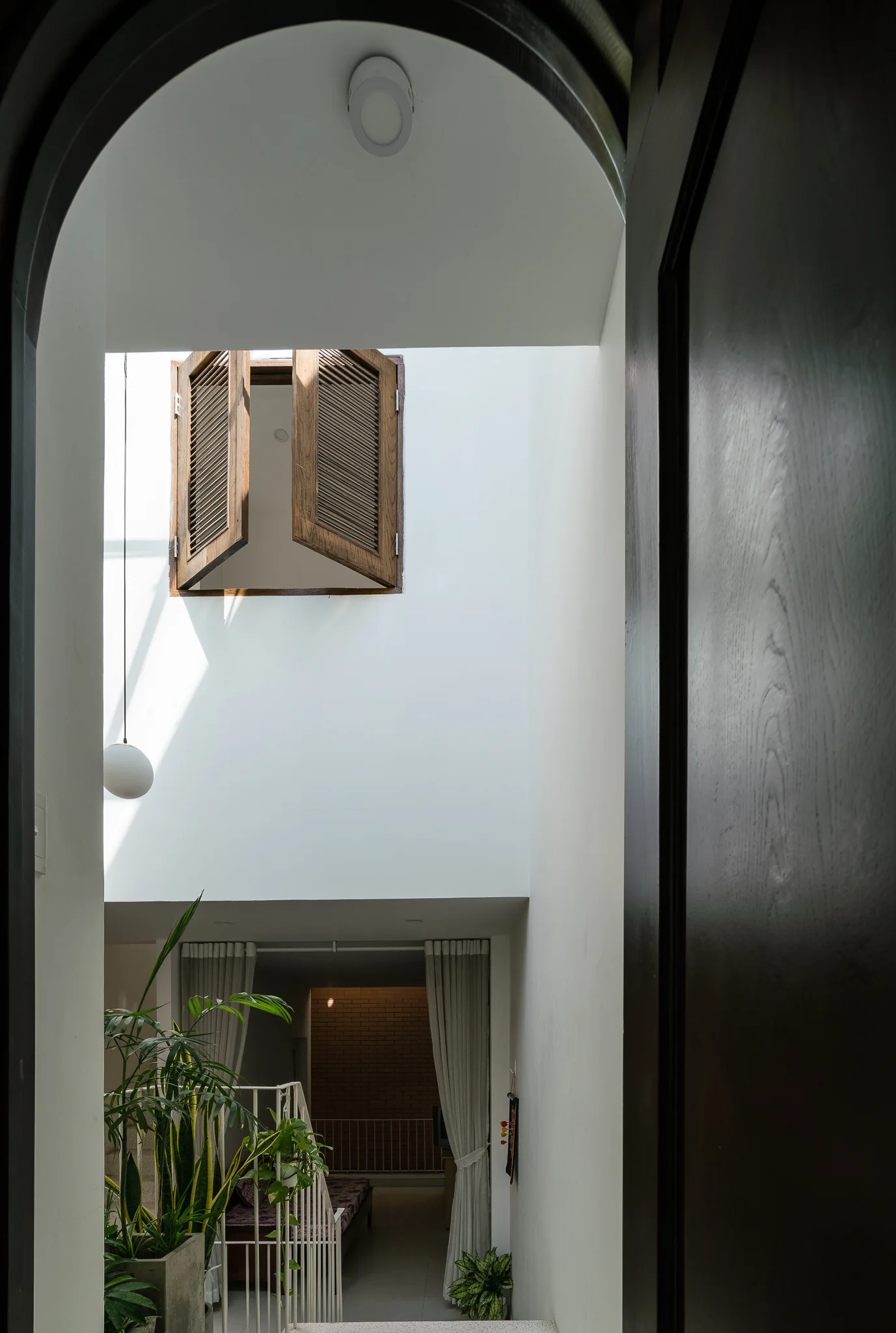 An arched doorway frames a multi-level white atrium featuring open wooden shutters on a high window, a staircase with metal railings, and potted plants.