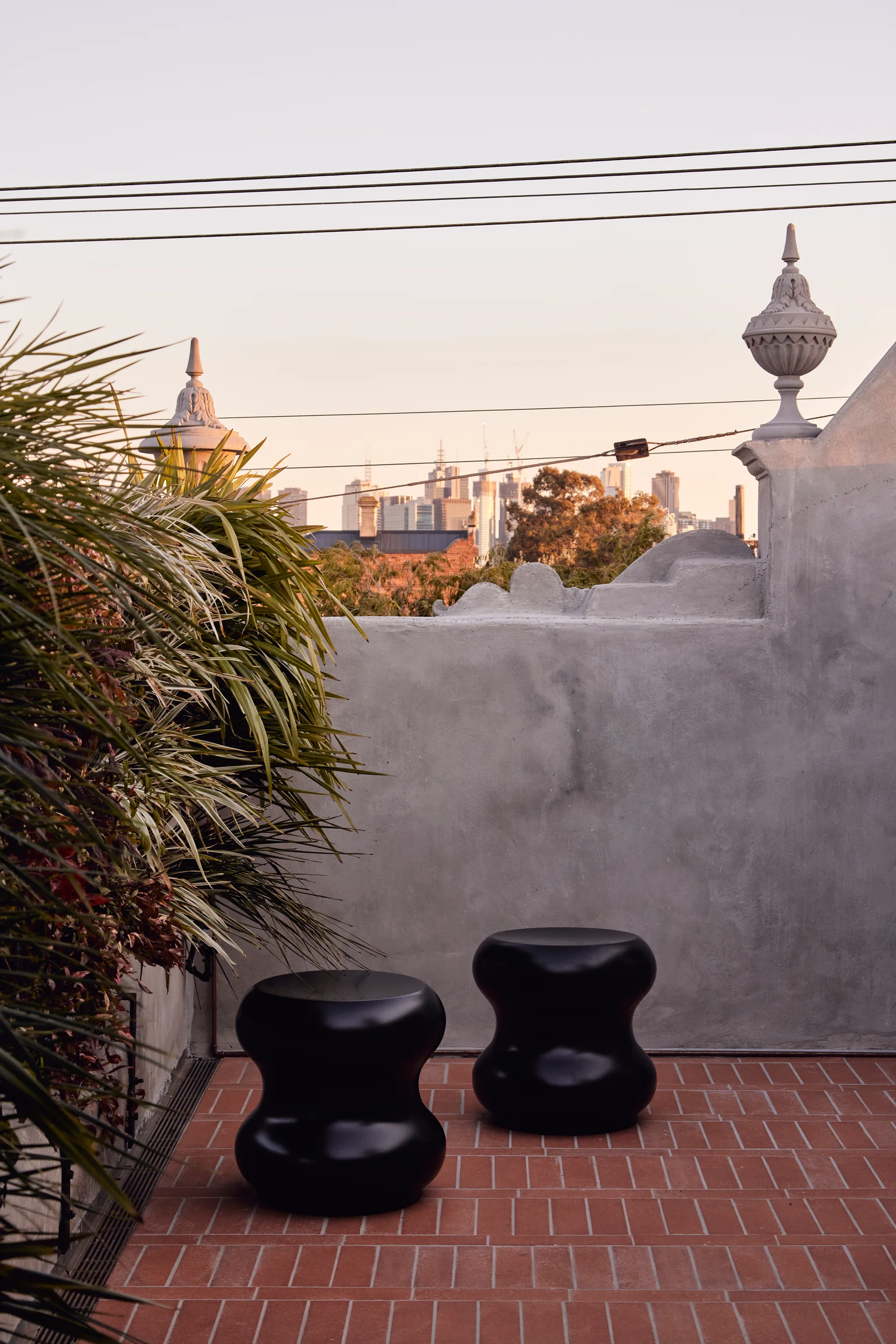 Two black sculptural stools sit on a terracotta tiled rooftop terrace against a grey concrete wall with decorative finials.
