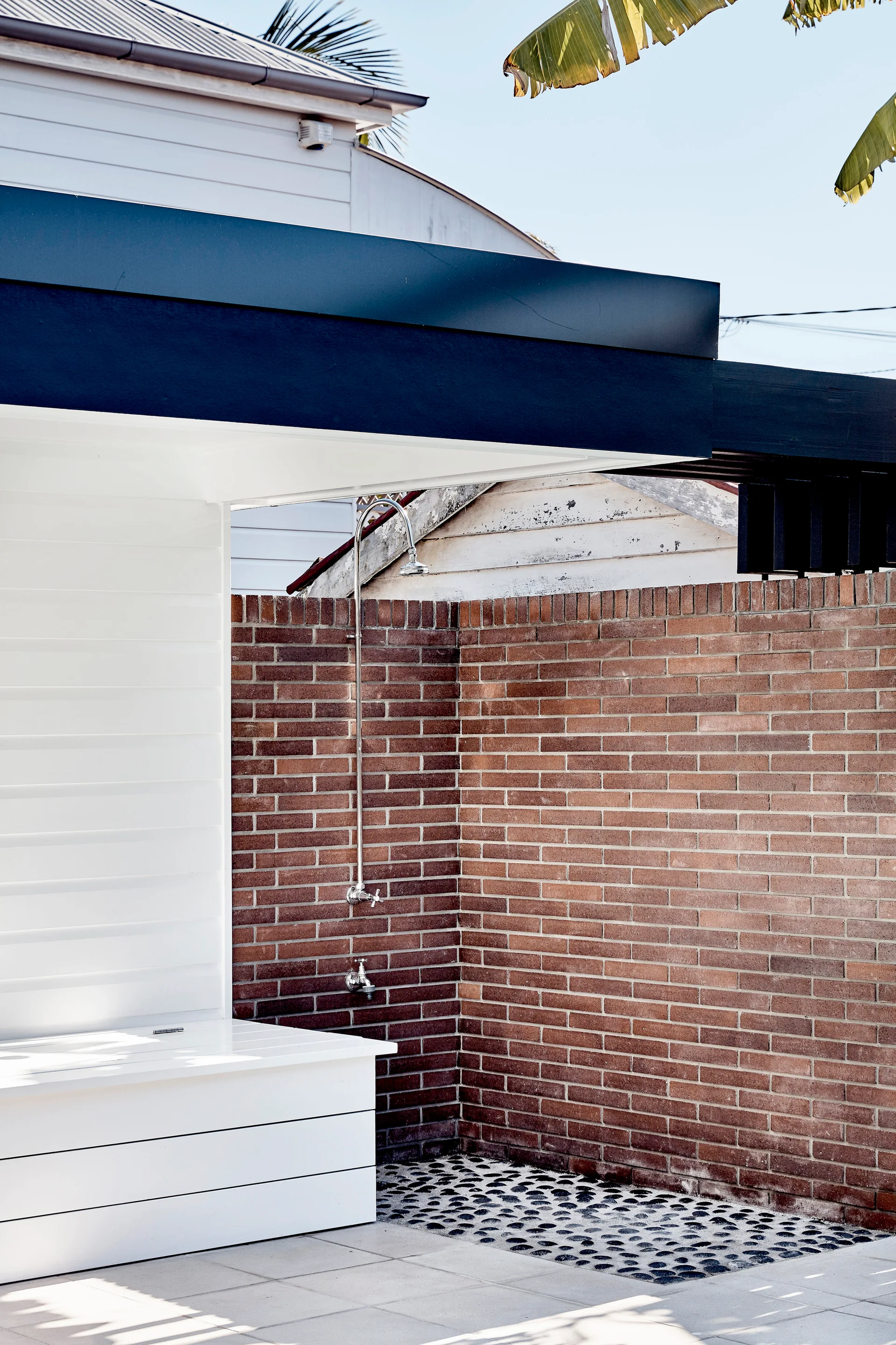 An outdoor chrome shower is mounted on a red brick wall next to a white wooden bench and a dark pebble floor inlay.