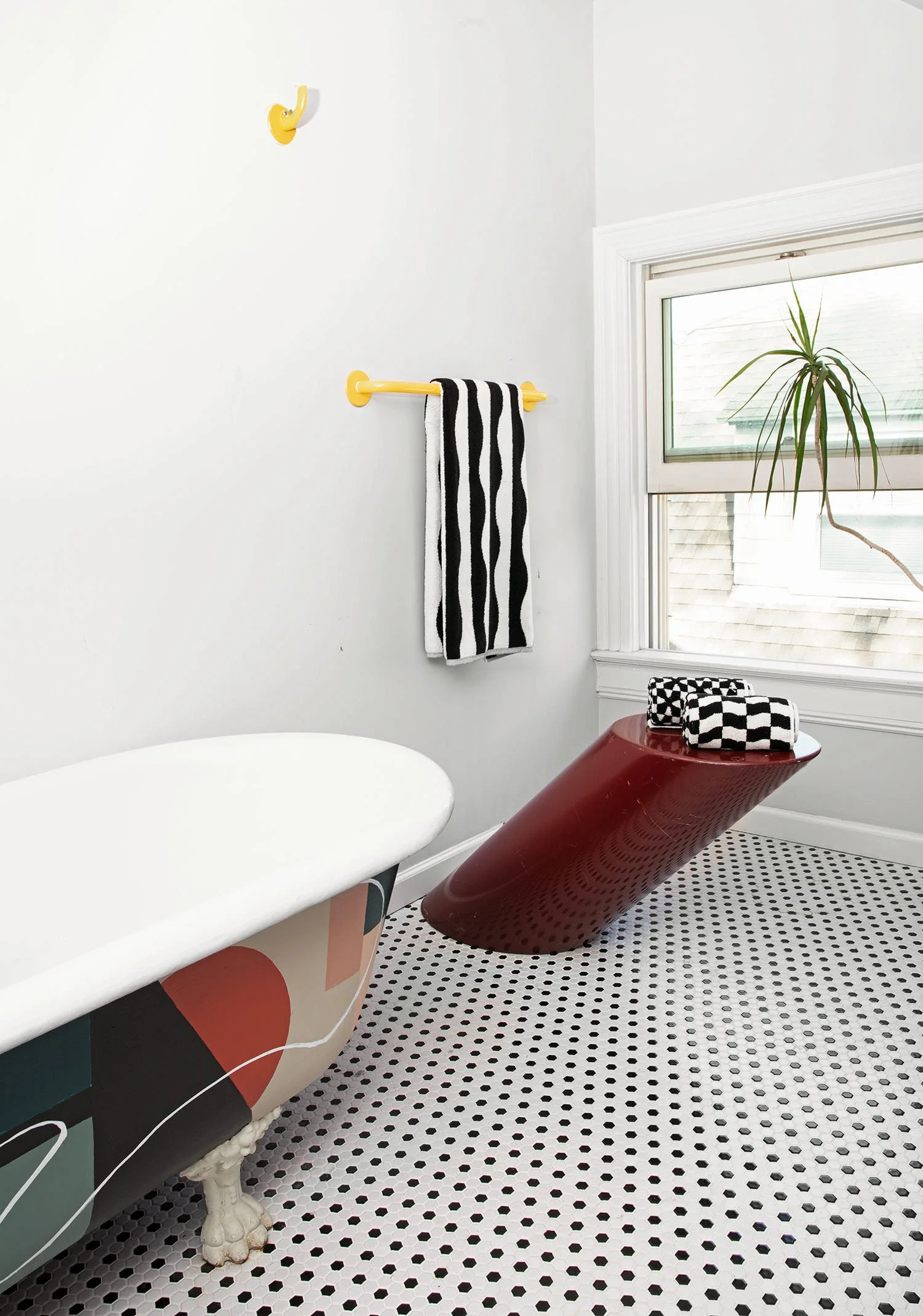 A bright bathroom features a graphic-patterned clawfoot tub, a slanted maroon side table, yellow wall fixtures, and black-and-white hex tile flooring.