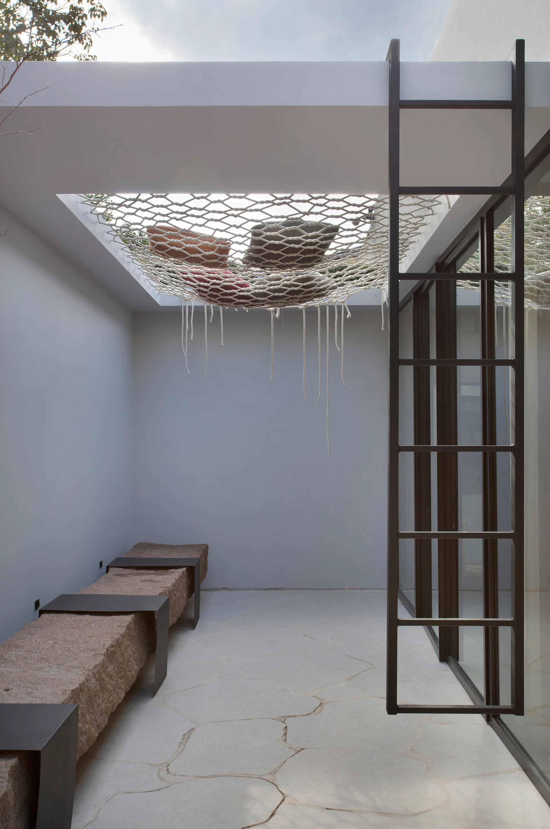 A minimalist outdoor patio features raw stone block benches, irregular flagstone flooring, and a woven rope netting bed suspended over a ceiling cutout.