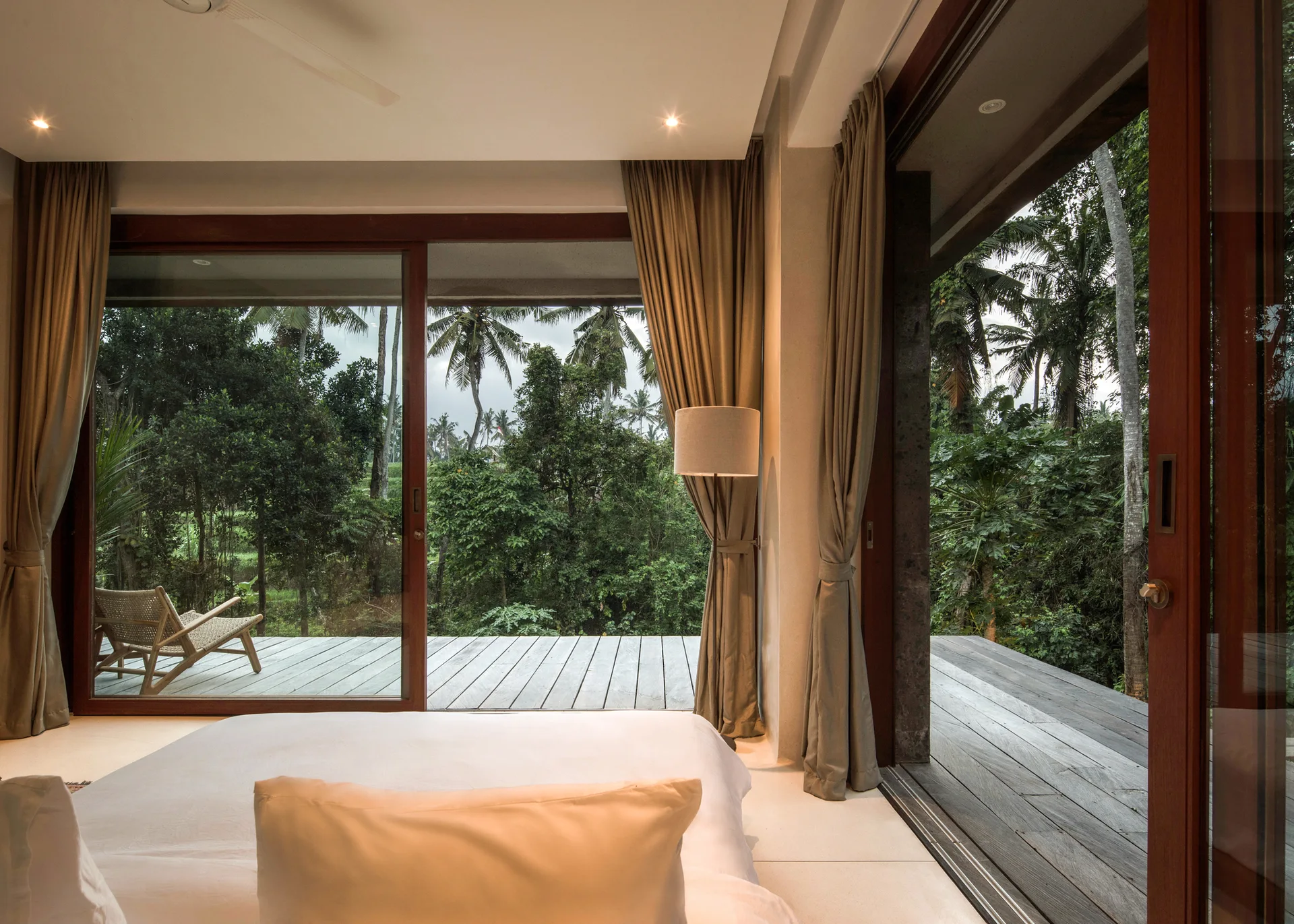 A minimalist bedroom with a white bed faces large wood-framed sliding glass doors that open onto a wooden deck overlooking a tropical forest.