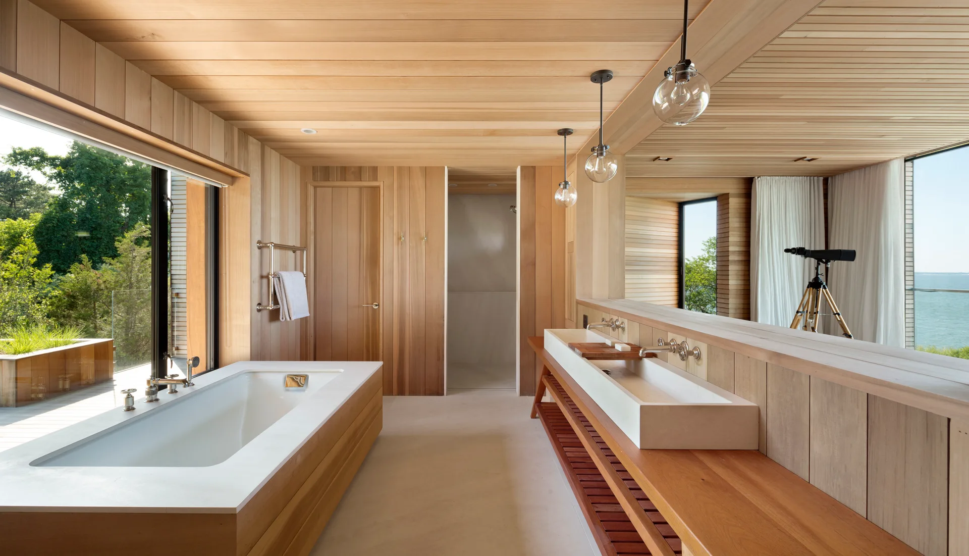 A minimalist wood-paneled bathroom features a white rectangular bathtub, a long wooden vanity with double sinks, and a large mirror.