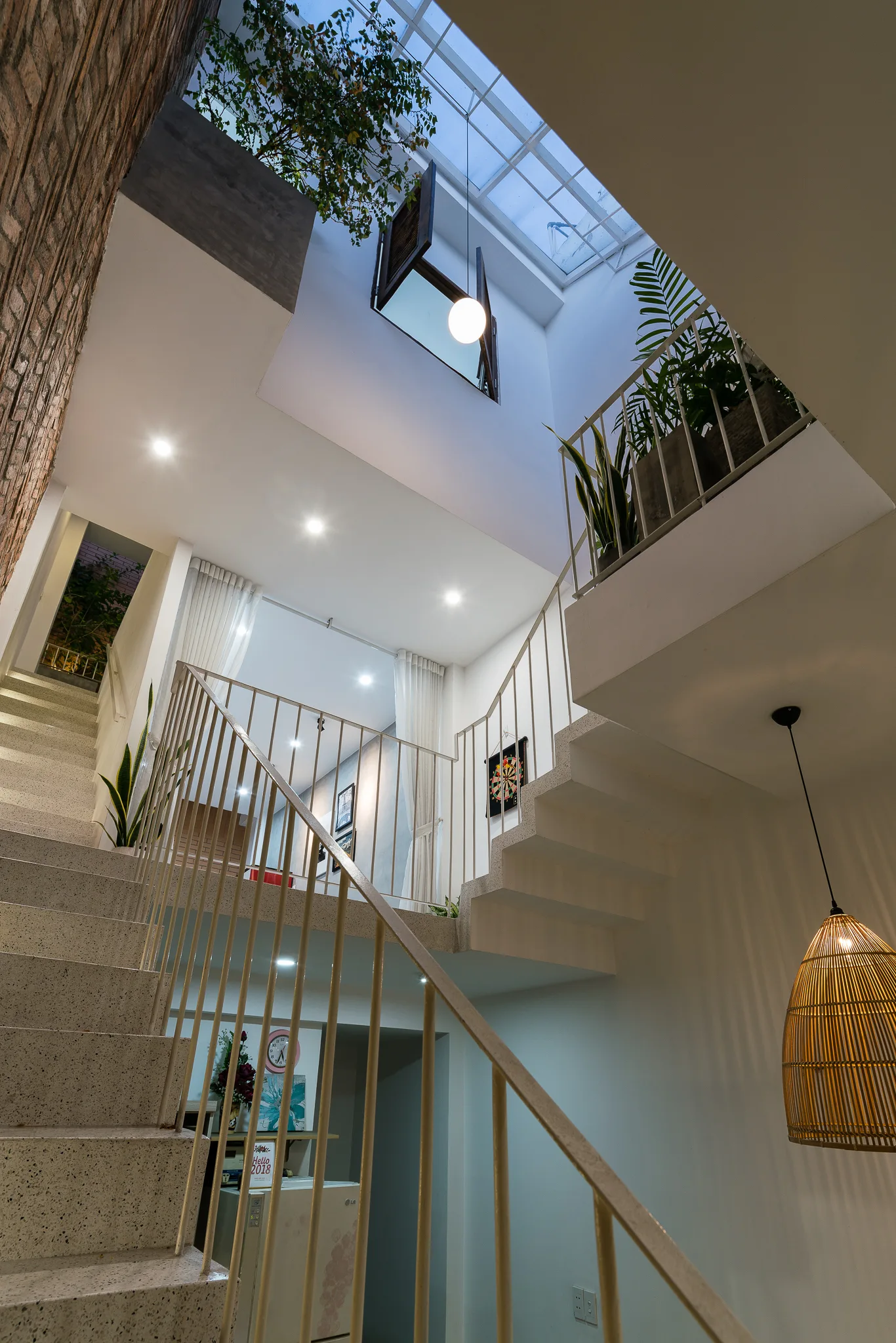 A multi-level terrazzo staircase features slender metal railings under a bright overhead skylight with green plants on the landings.