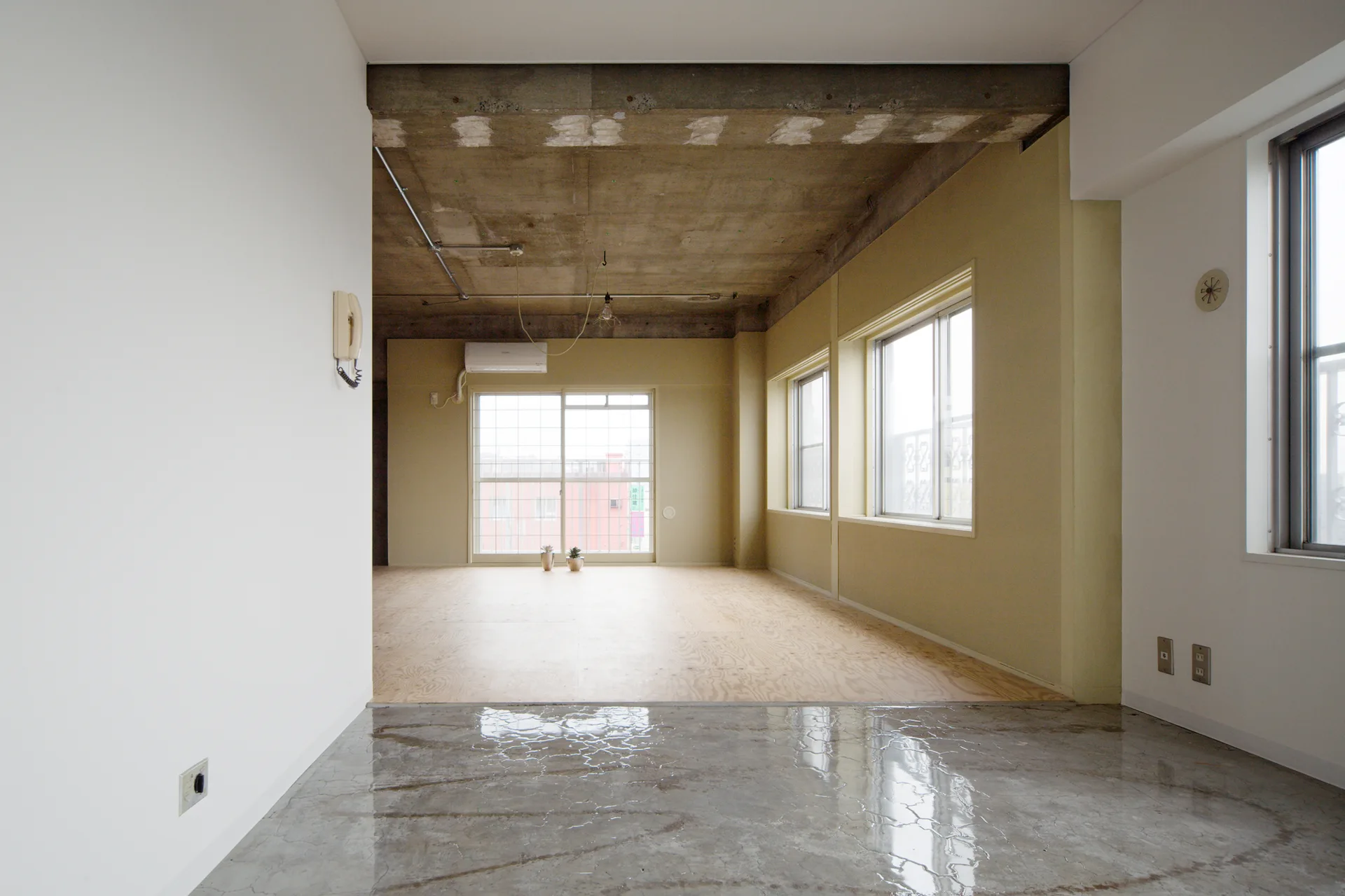 An empty industrial room features an exposed concrete ceiling, pale yellow walls, large windows, and a light tan plywood floor.
