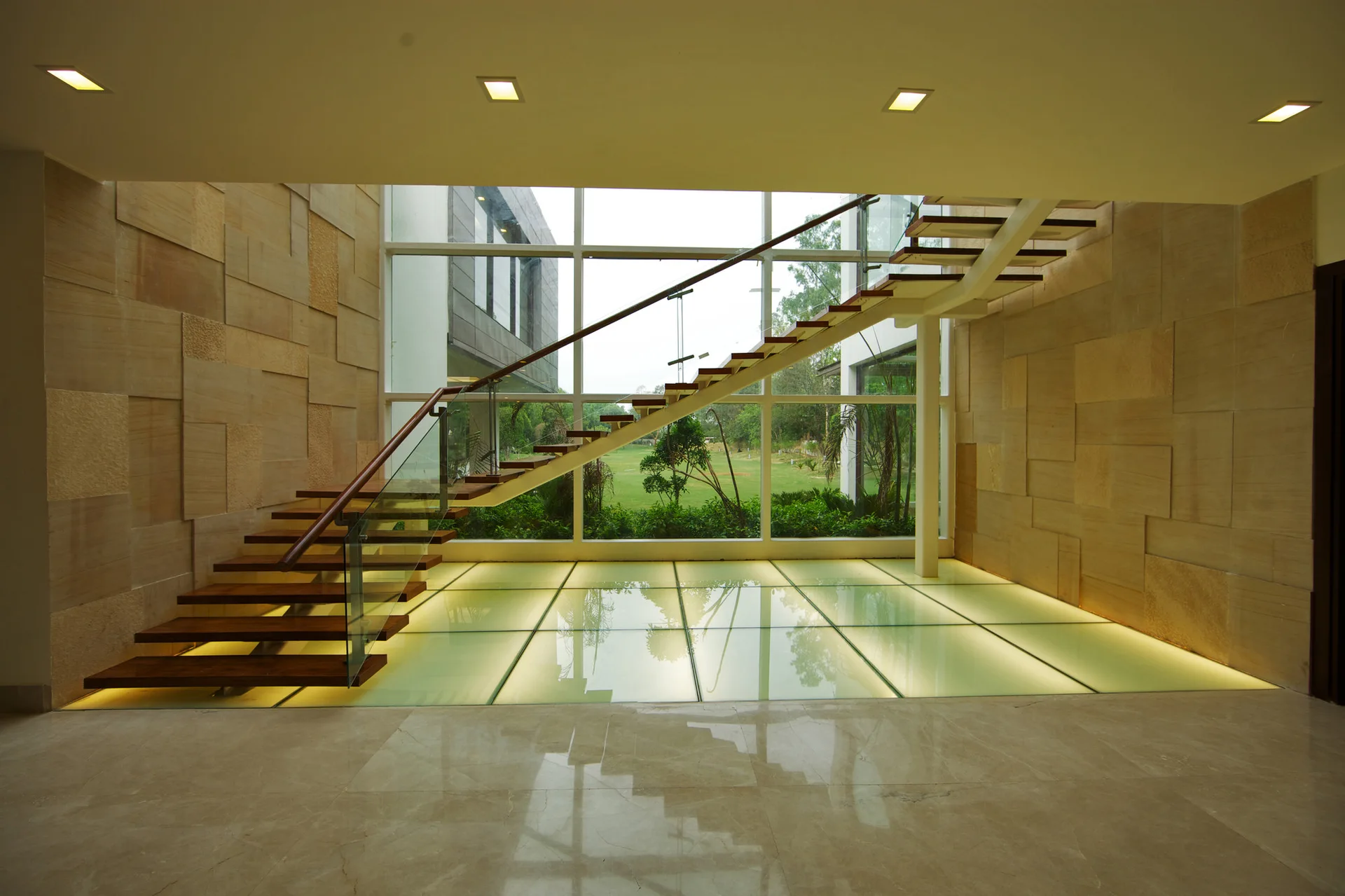 An open-riser wooden staircase with glass railings ascends next to a tall window wall over illuminated glass floor tiles and stone-clad walls.