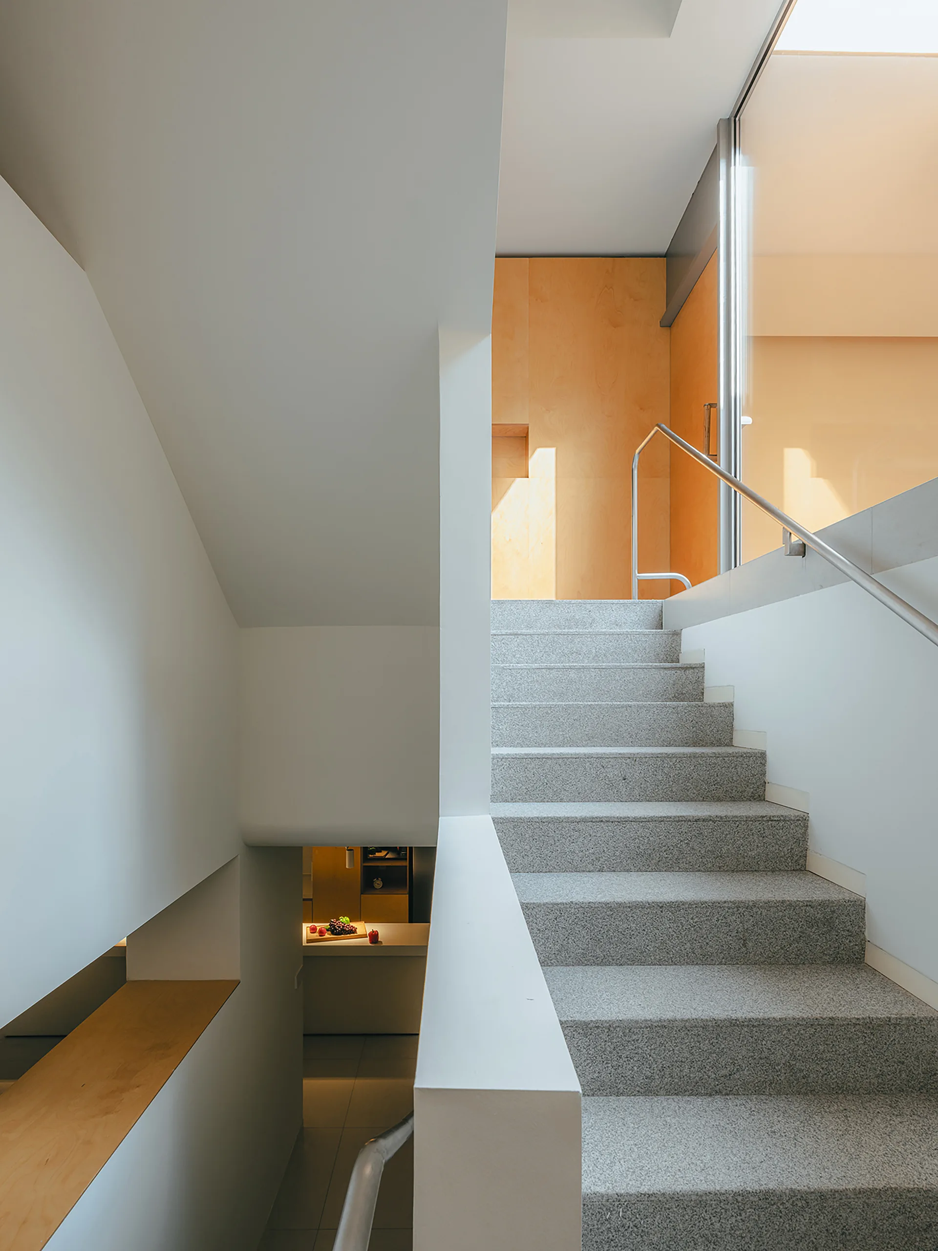 A grey speckled stone staircase rises between smooth white walls toward a sunlit landing with warm wood paneling.