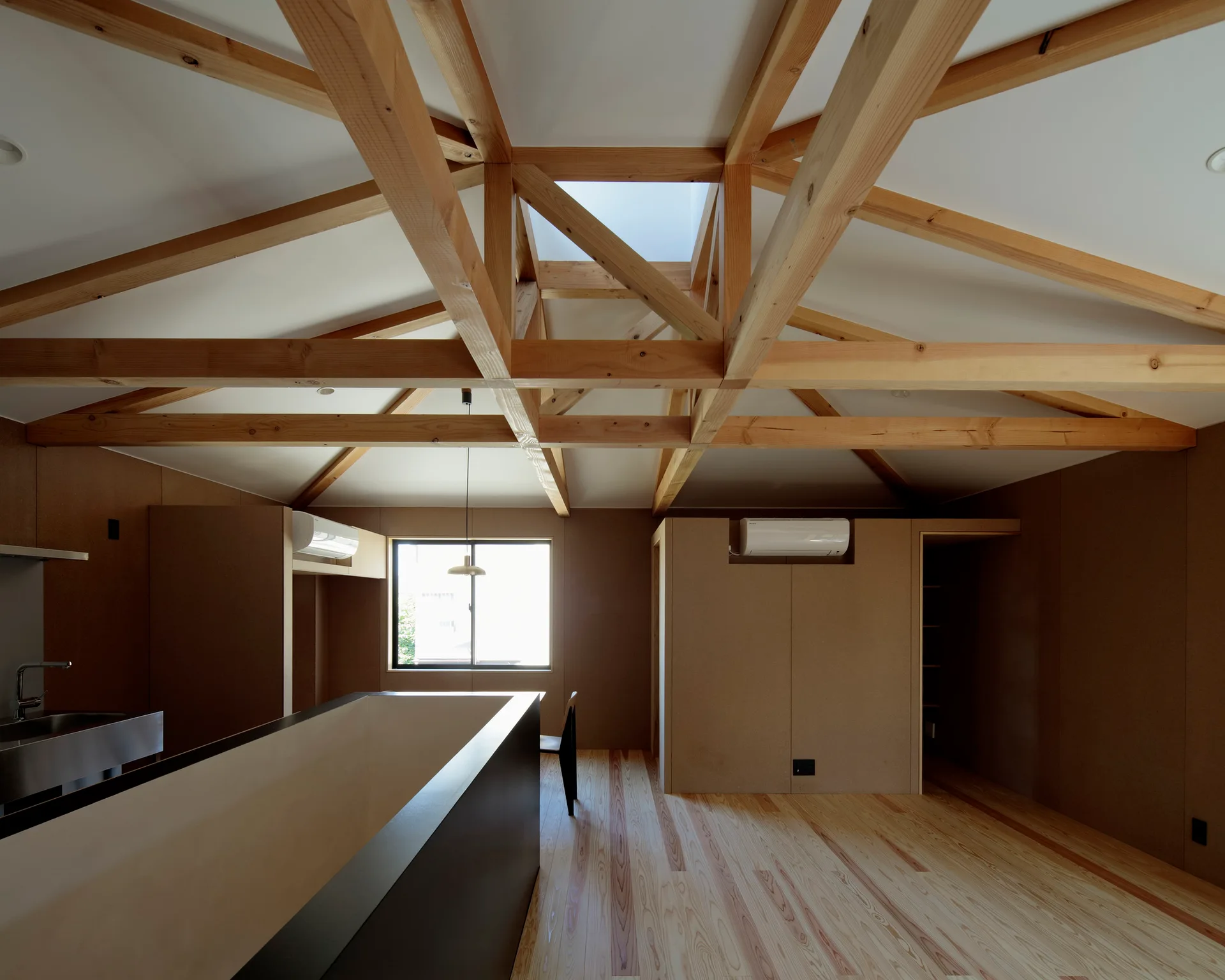 An open kitchen features a vaulted white ceiling with exposed timber trusses, a skylight, light wood floors, and tan wall panels.