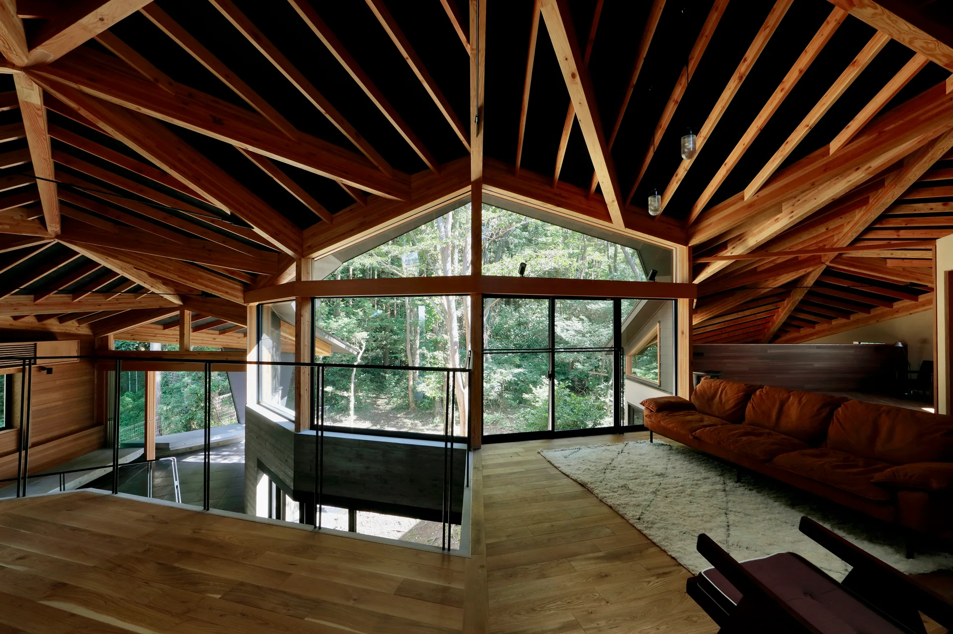 A modern loft living area featuring a complex exposed timber vaulted ceiling, a brown sofa, and large windows looking out to a dense green forest.