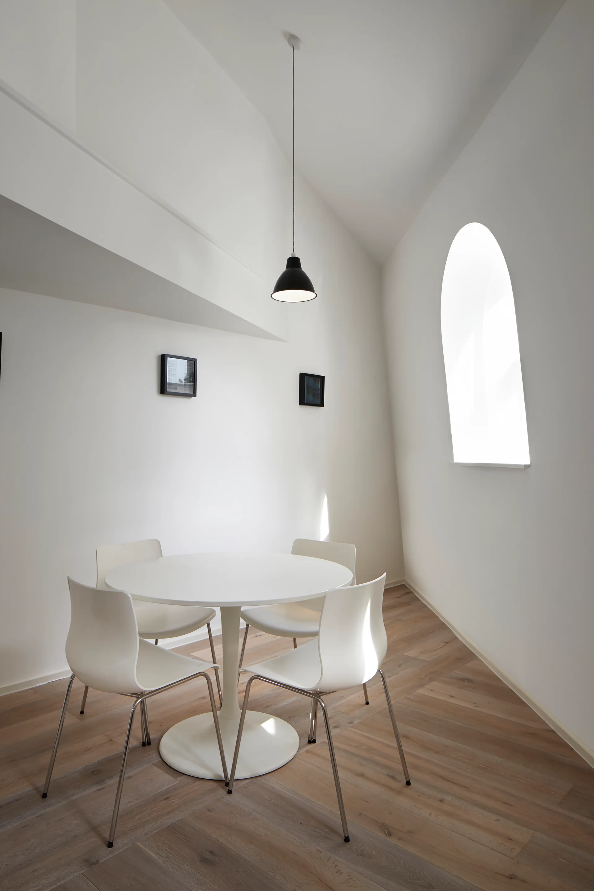 A white pedestal dining table and four matching chairs sit on light wood floors beneath a black pendant light and next to an arched window.