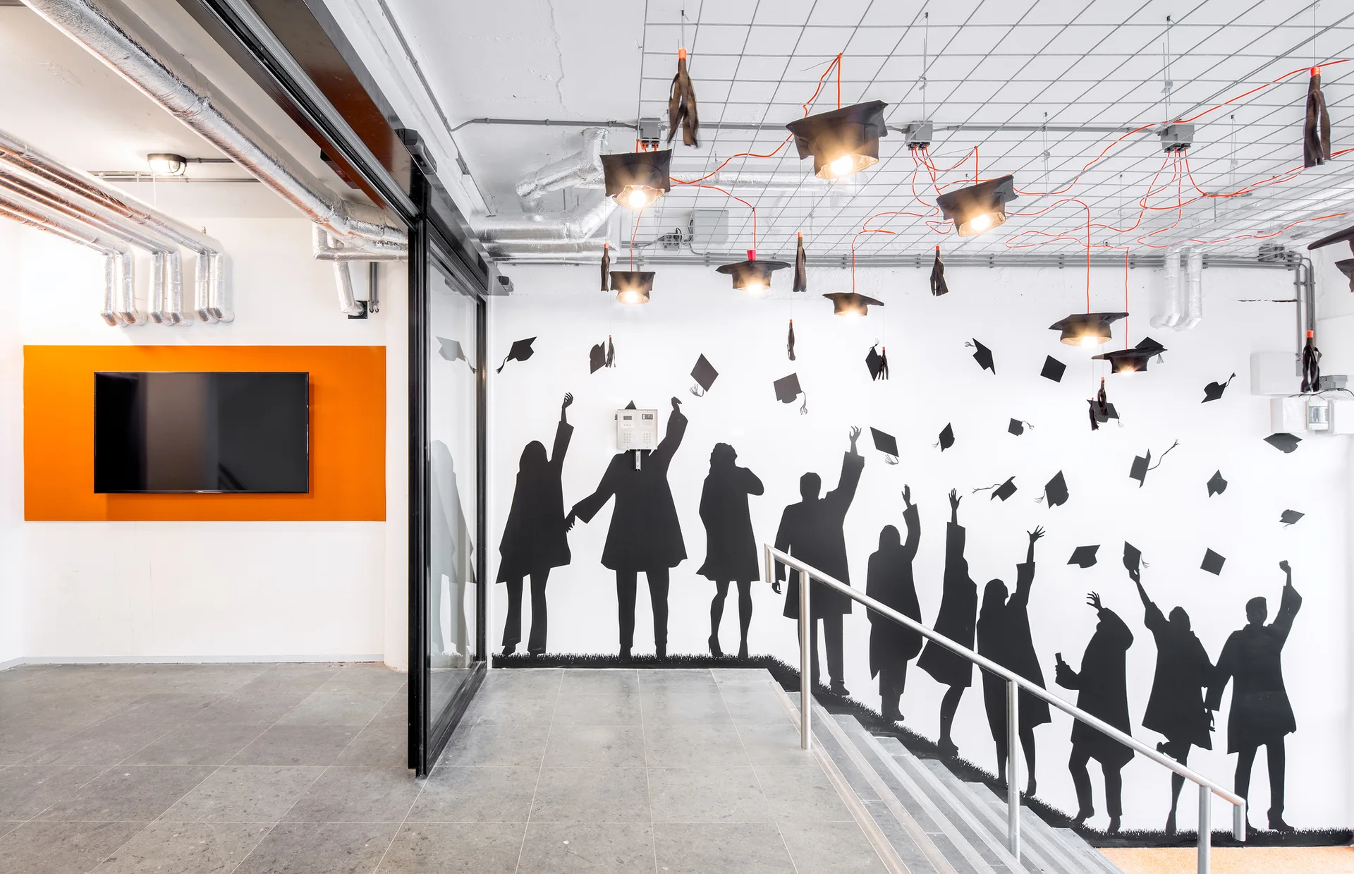 An industrial staircase features a black-and-white silhouette mural of graduates, concrete tile flooring, and graduation-cap pendant lights.