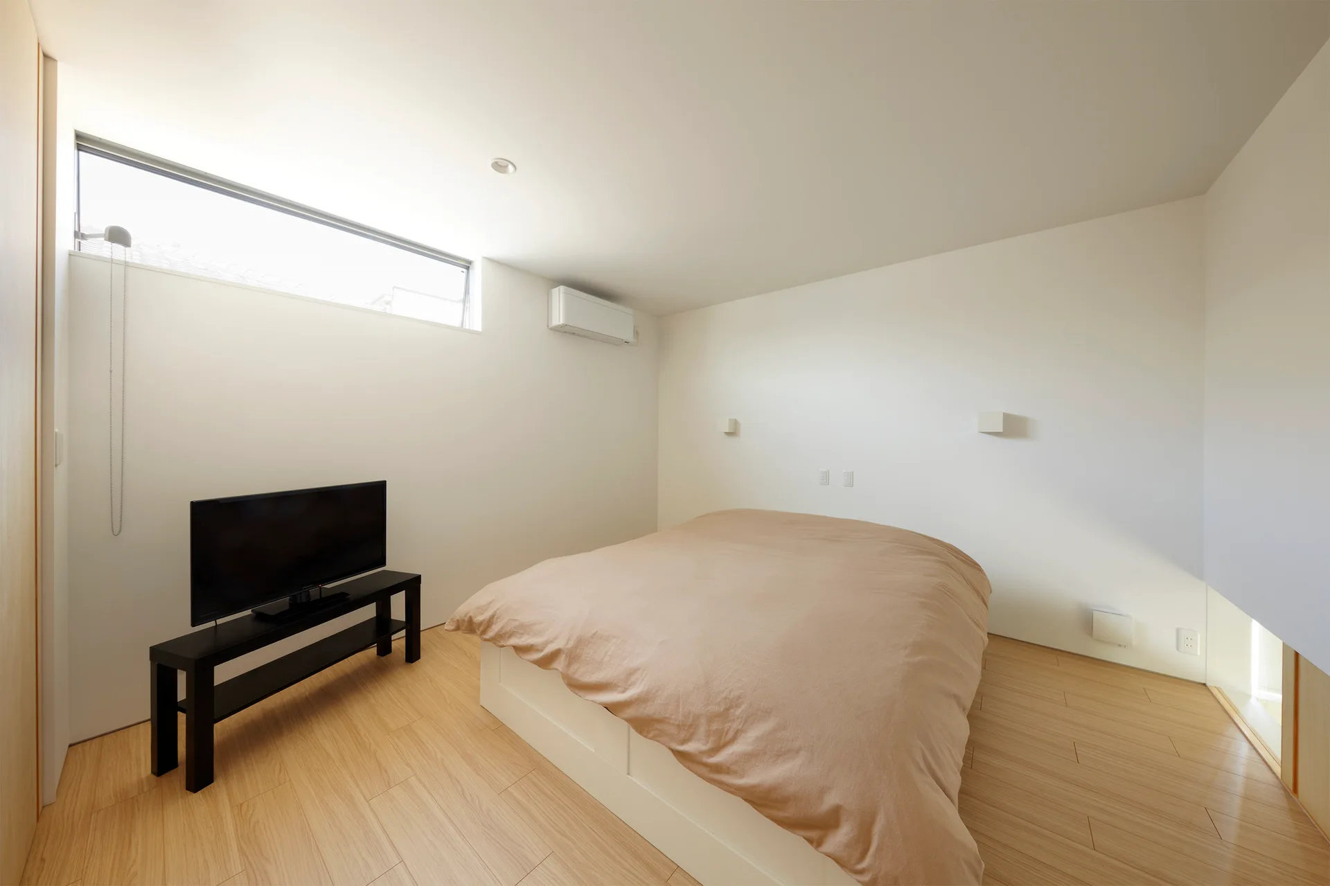 A minimalist bedroom featuring a bed with beige bedding, a flat-screen television on a black stand, white walls, and a high horizontal window.