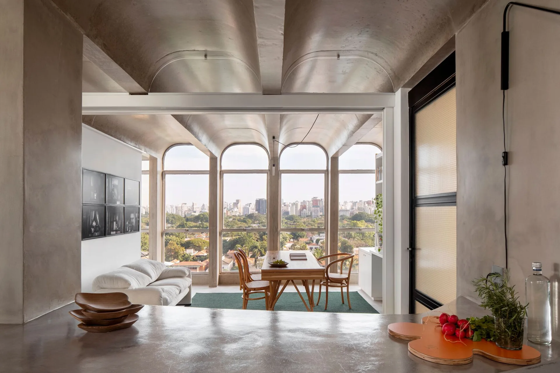 A dining table sits under a vaulted concrete ceiling next to large arched windows looking out at a city skyline.