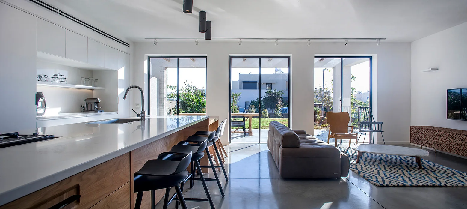 An open-plan kitchen and living area features a wood island with black stools, polished concrete flooring, and sliding glass doors leading to a garden.