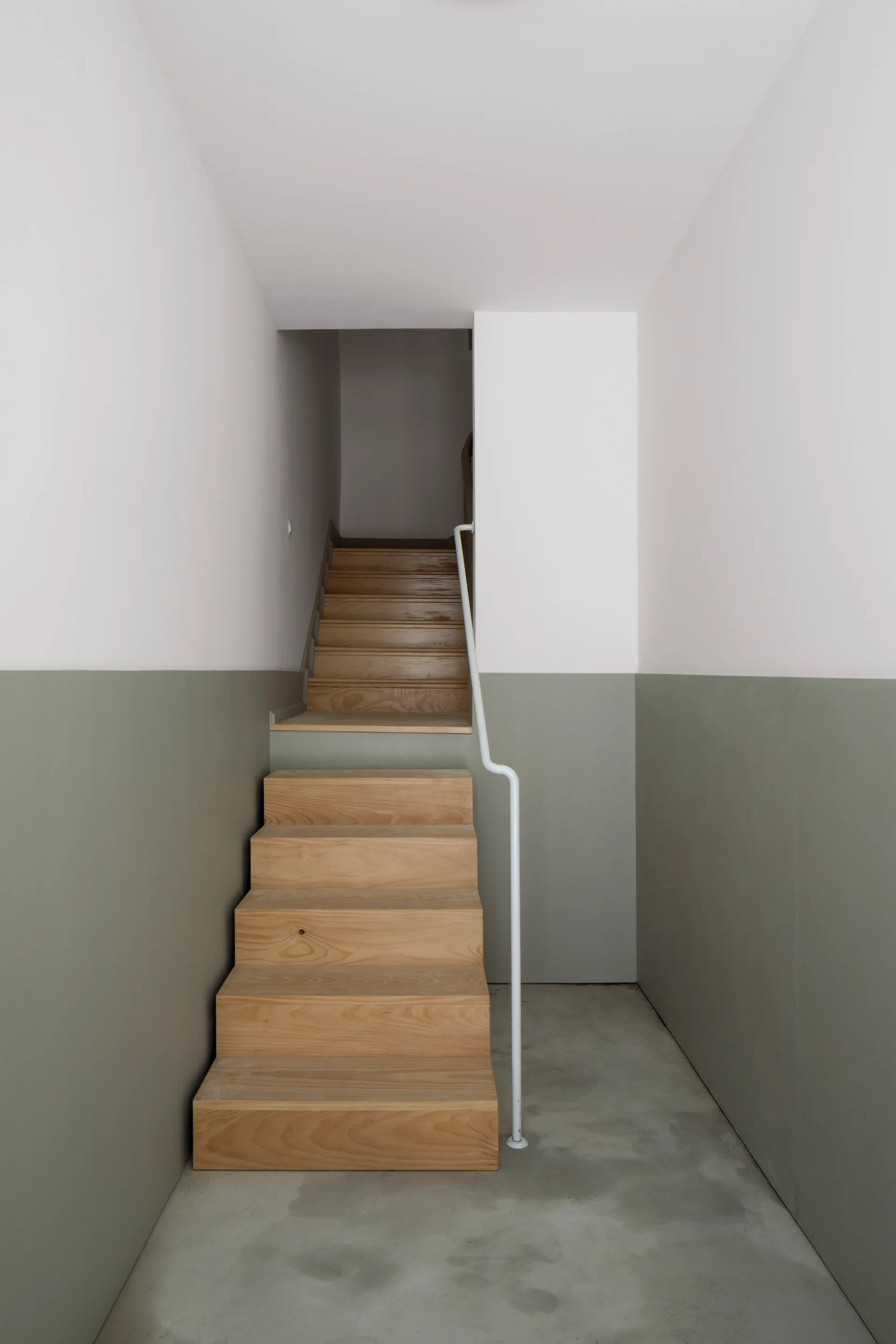 A wooden staircase with a thin white metal handrail sits between walls painted sage green on the bottom and white on top.