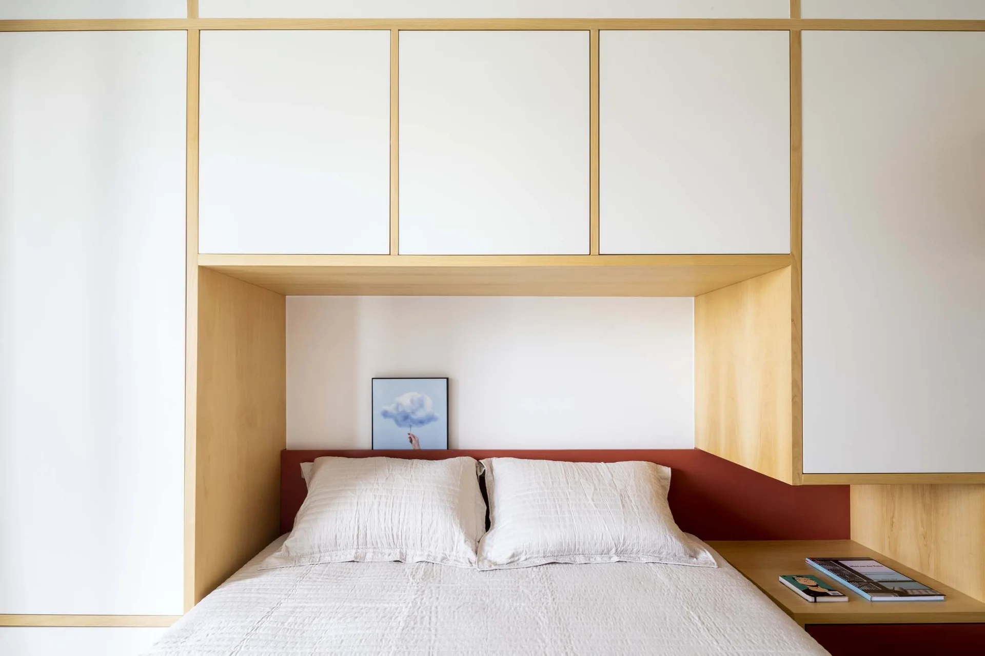 A double bed with light bedding is recessed into a built-in white and wood storage system with a red accent panel behind the pillows.