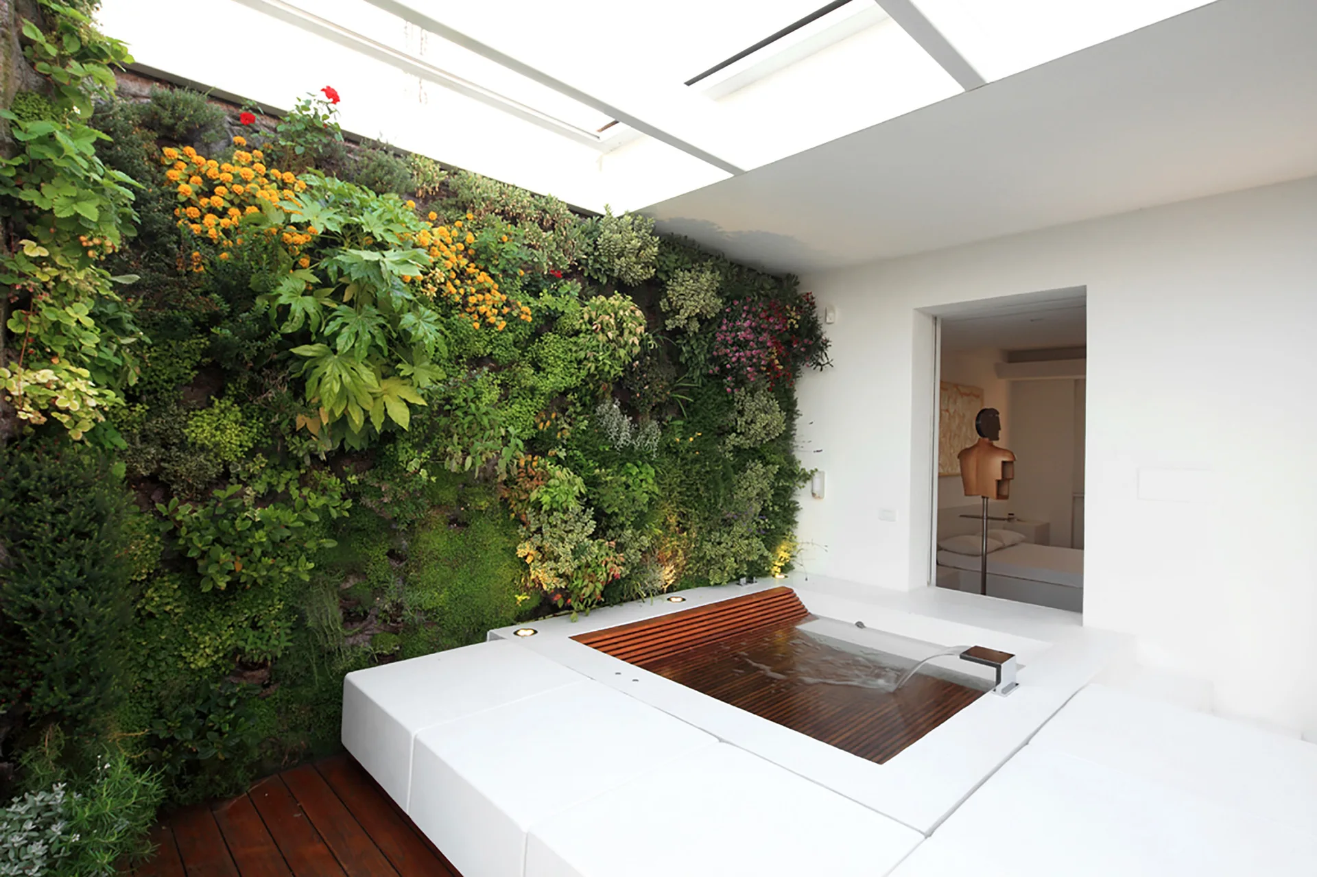 A sunken spa pool with a wooden slatted cover is set beside a massive vertical garden wall of lush green plants under a bright skylight.