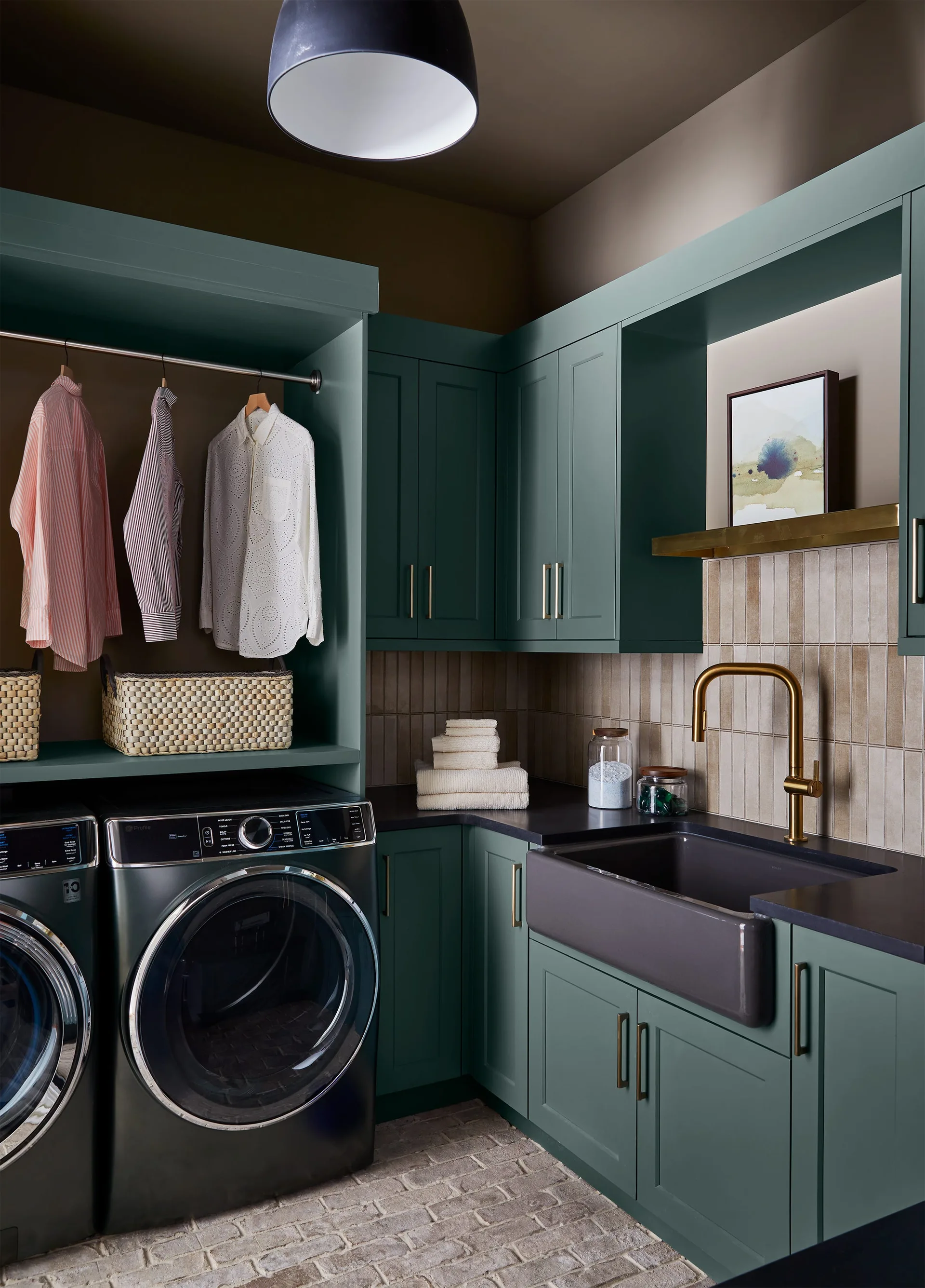 A laundry room featuring dark green built-in cabinetry, a charcoal farmhouse sink with a gold faucet, and front-loading washer and dryer units.