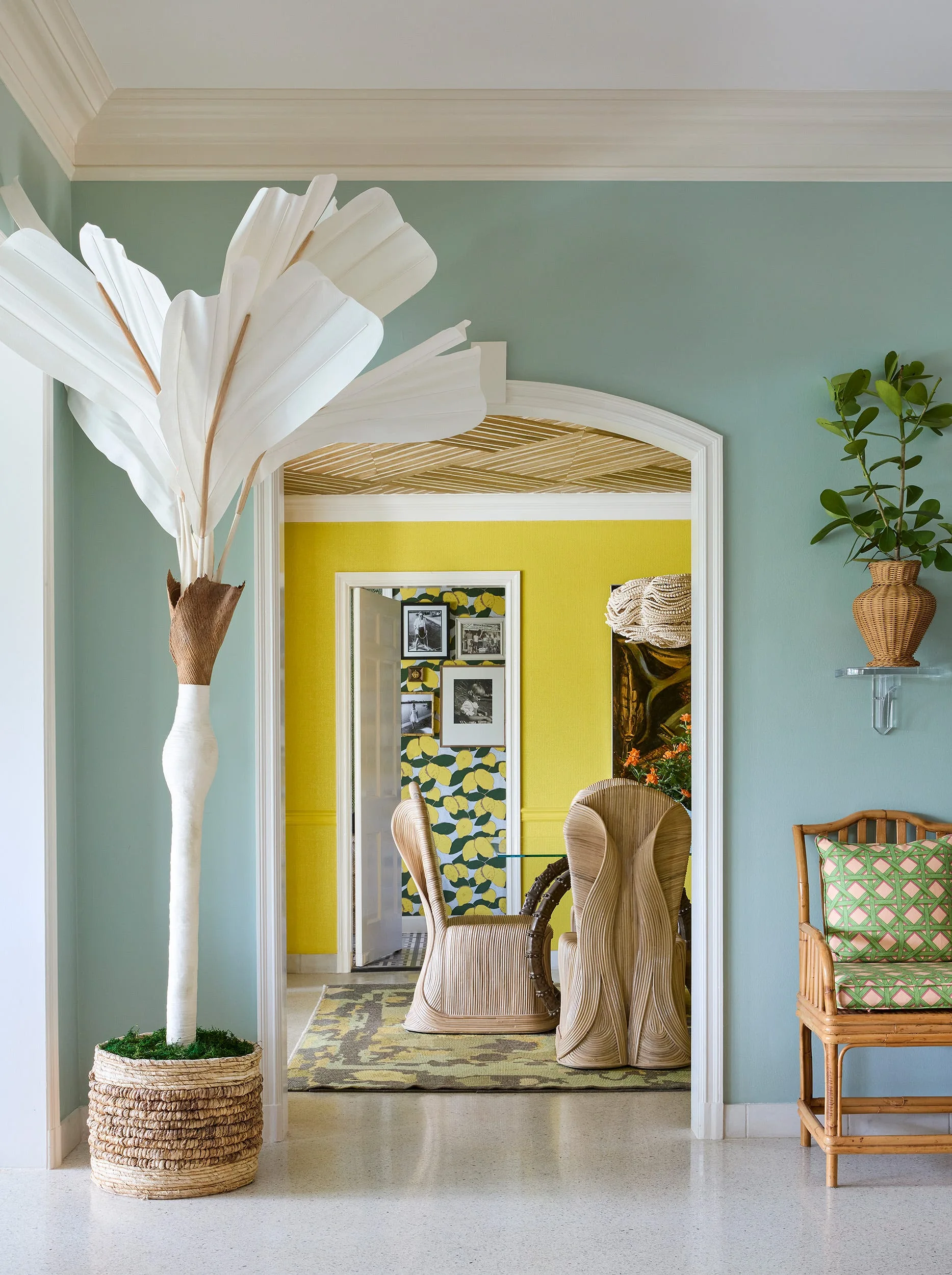 An arched doorway links a light blue room to a bright yellow dining space featuring sculptural rattan chairs and lemon-print wallpaper.