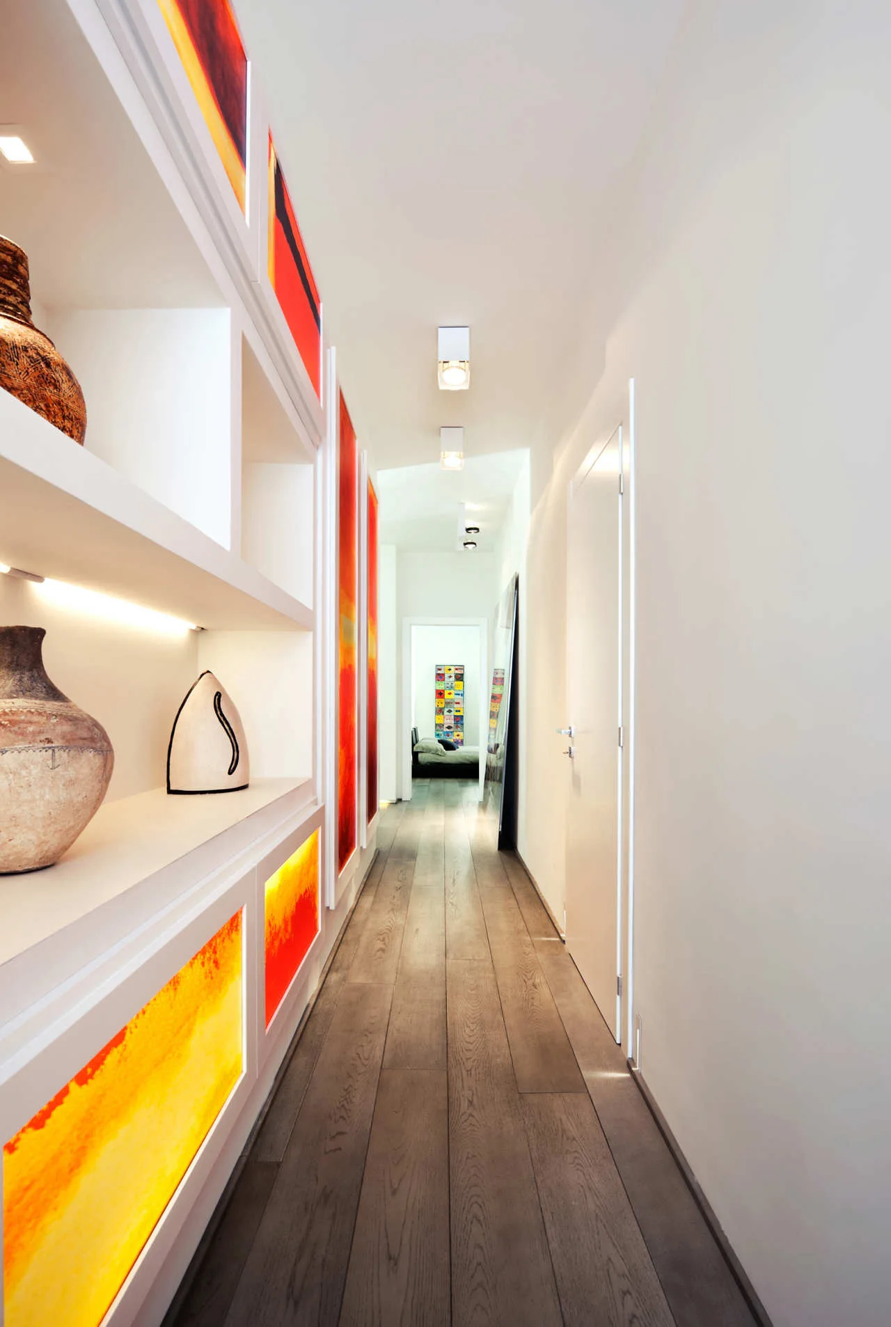 A long white hallway with dark wood flooring features a built-in shelving unit illuminated by warm orange backlit panels and recessed ceiling lights.