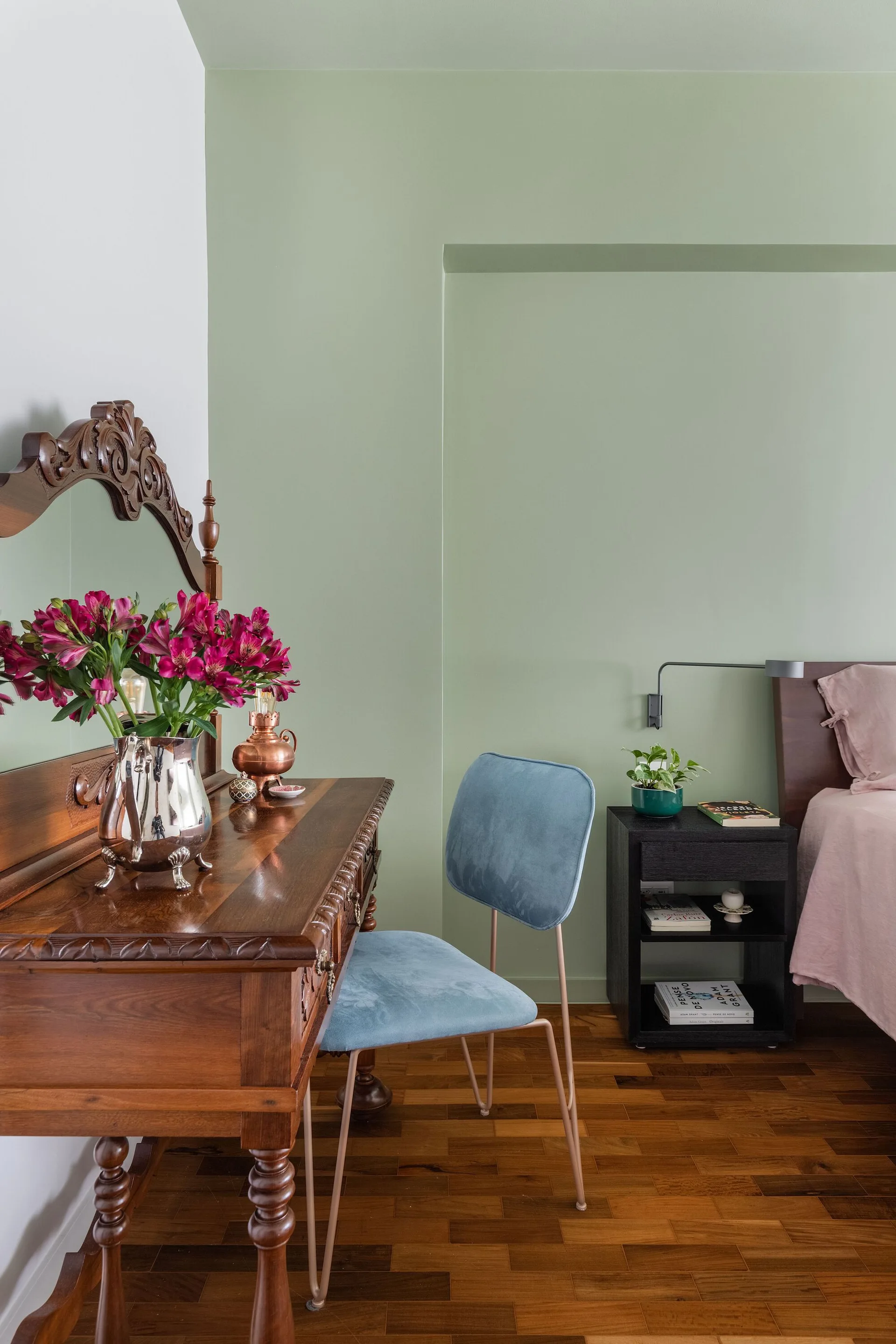 A bedroom corner with sage green walls features a carved wood vanity, a blue velvet chair, and a black nightstand next to a bed.