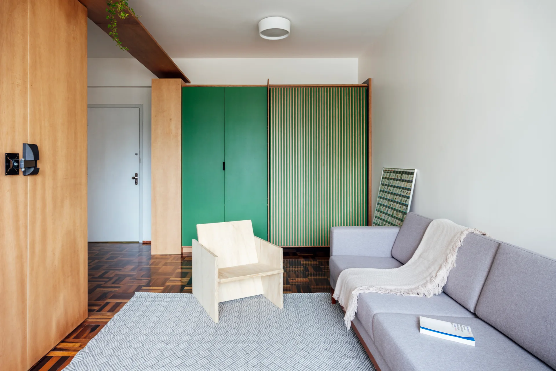 A modern living room featuring a grey sofa, a light wood geometric lounge chair, parquet flooring, and a forest green wardrobe with wood slats.