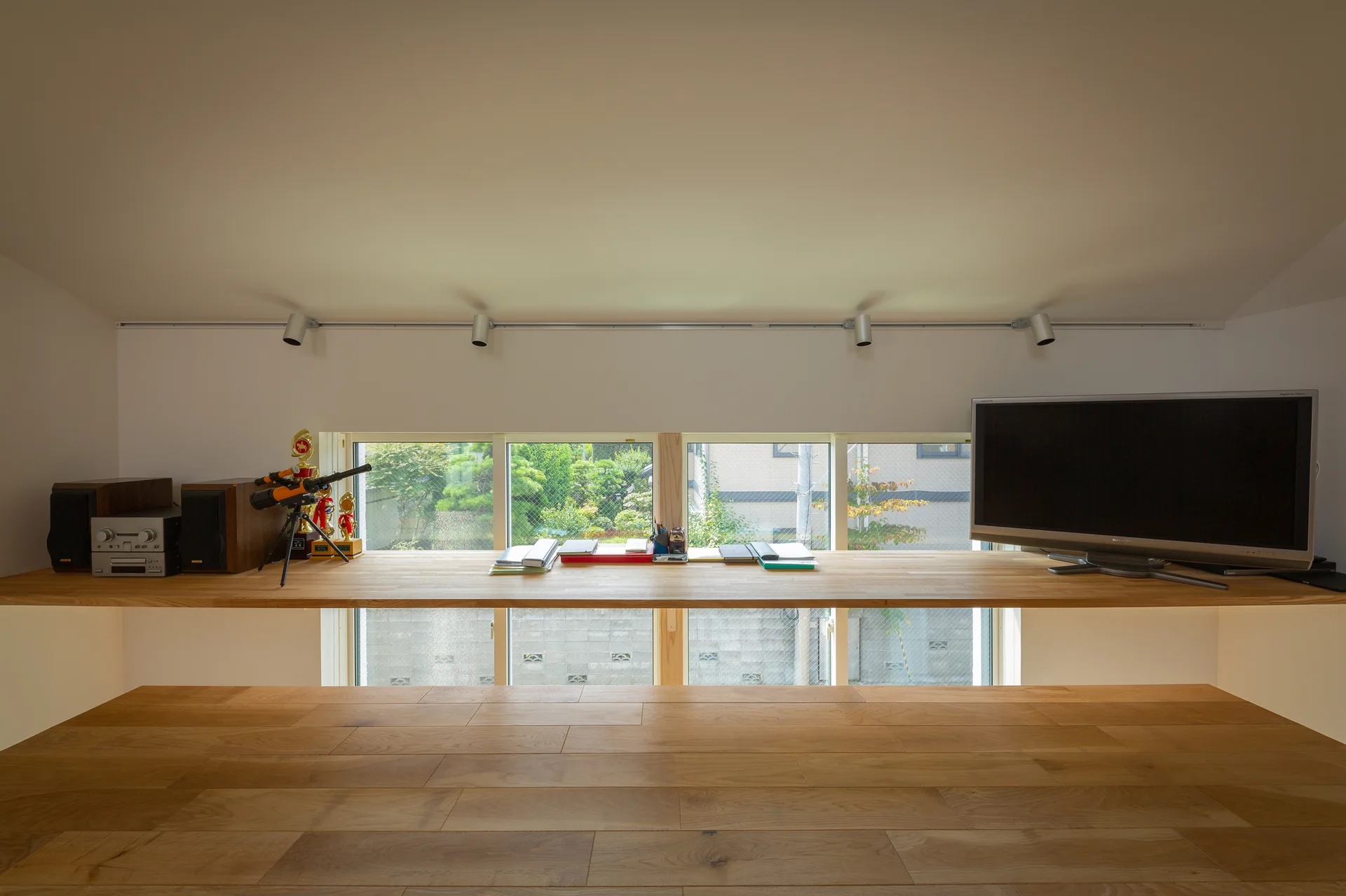 A minimalist wooden desk spans a low horizontal window beneath a sloped white ceiling with track lighting in a home office study.