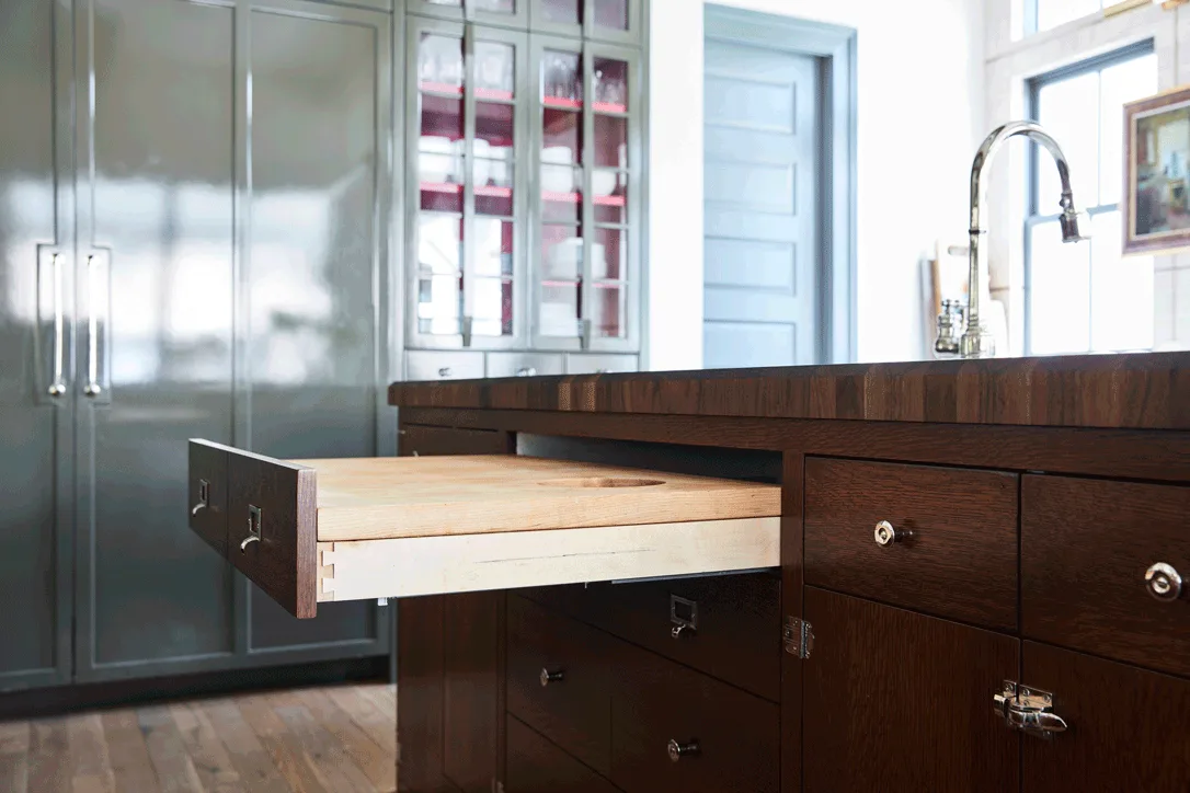 A close-up of a dark brown wood kitchen island showing an open drawer with an integrated pull-out cutting board, with grey cabinetry in the background.