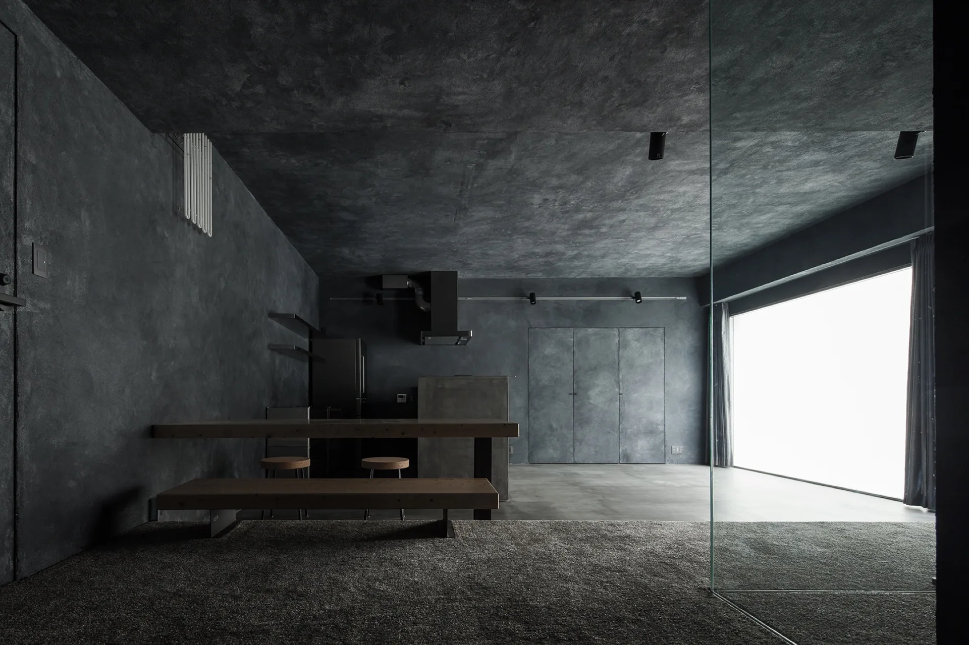 A minimalist kitchen and dining space with charcoal grey plaster walls, a wooden dining table, and a large bright window.