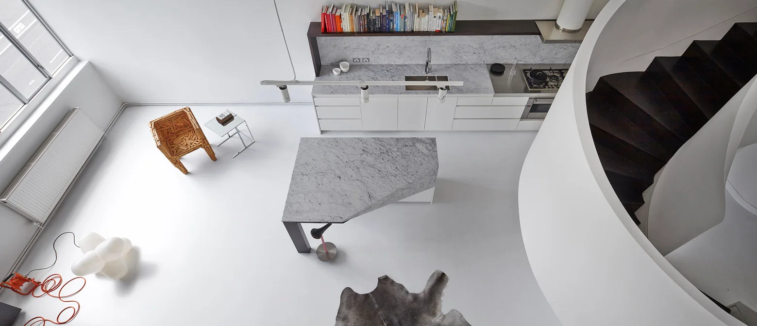 High-angle view of a minimalist kitchen with a trapezoidal marble island, seamless white floors, a dark curved staircase, and white cabinets.