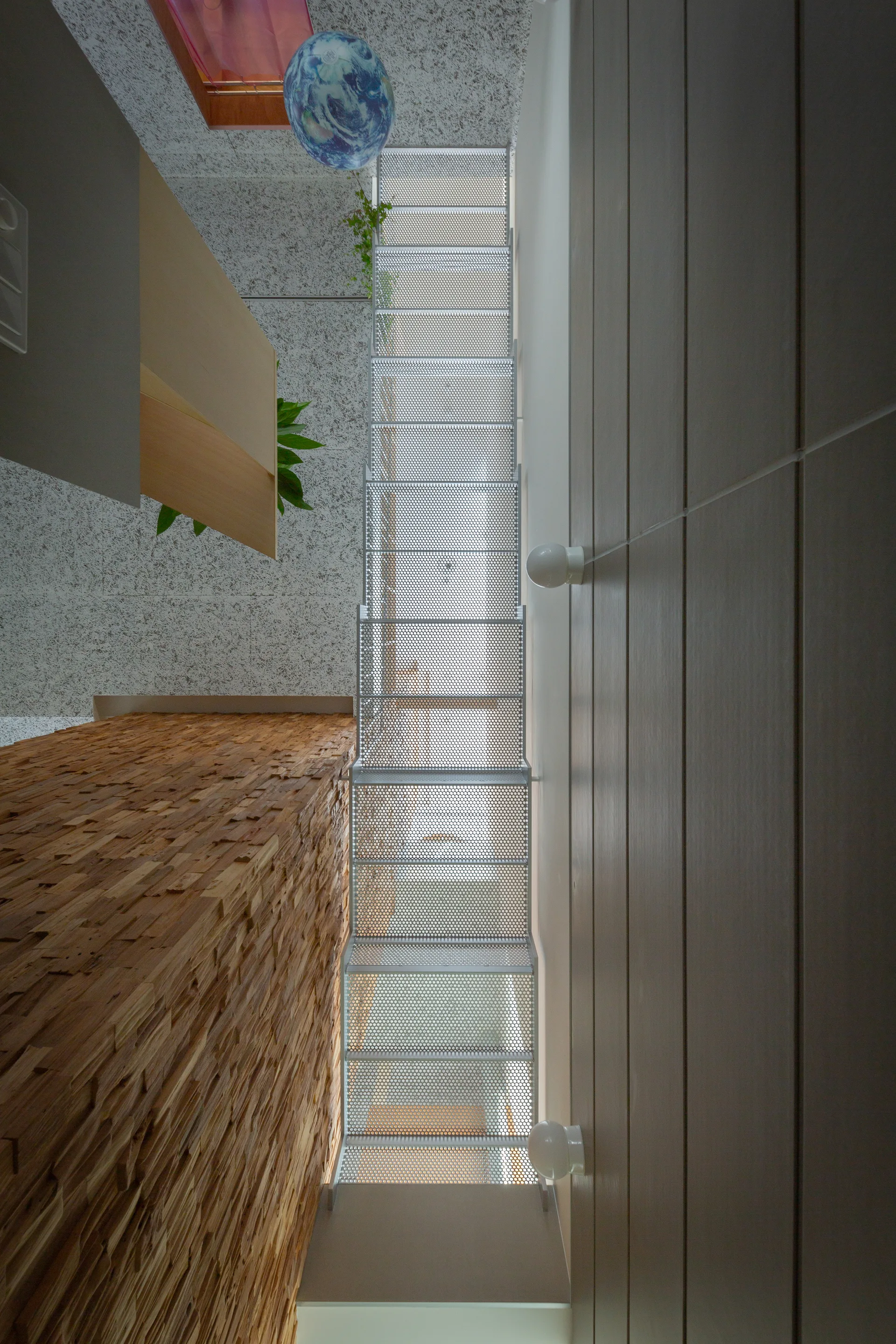 Looking up a narrow stairwell with a perforated metal staircase between a highly textured split-wood wall and a grey paneled wall.