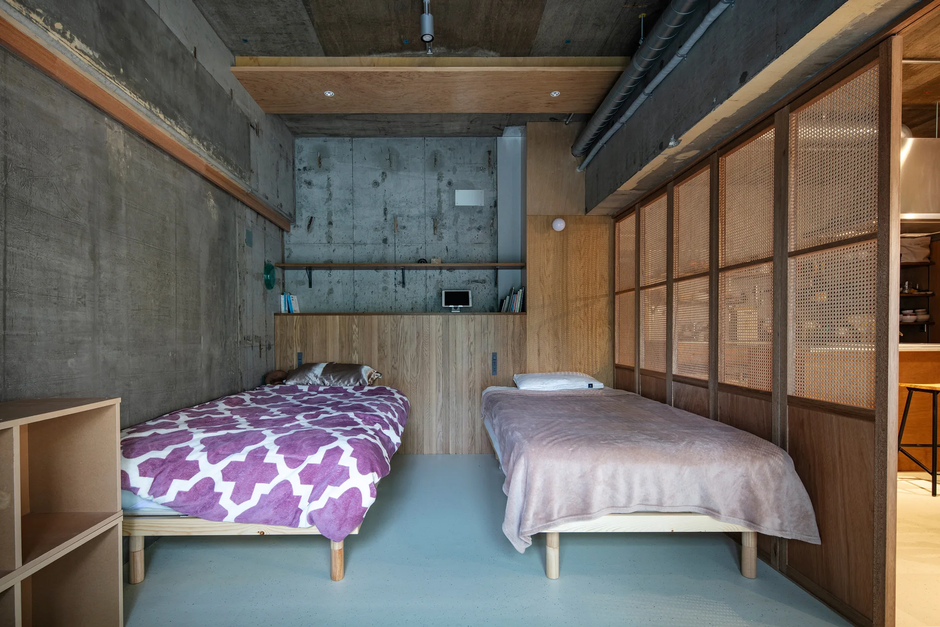 Two twin beds in an industrial room with raw concrete walls, a wood panel headboard, and a woven cane room divider.