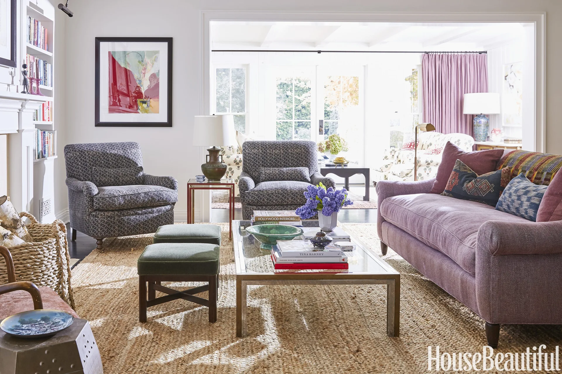 A bright living room features a plum-colored sofa, two patterned gray armchairs, two green stools, and a glass coffee table on a large jute rug.
