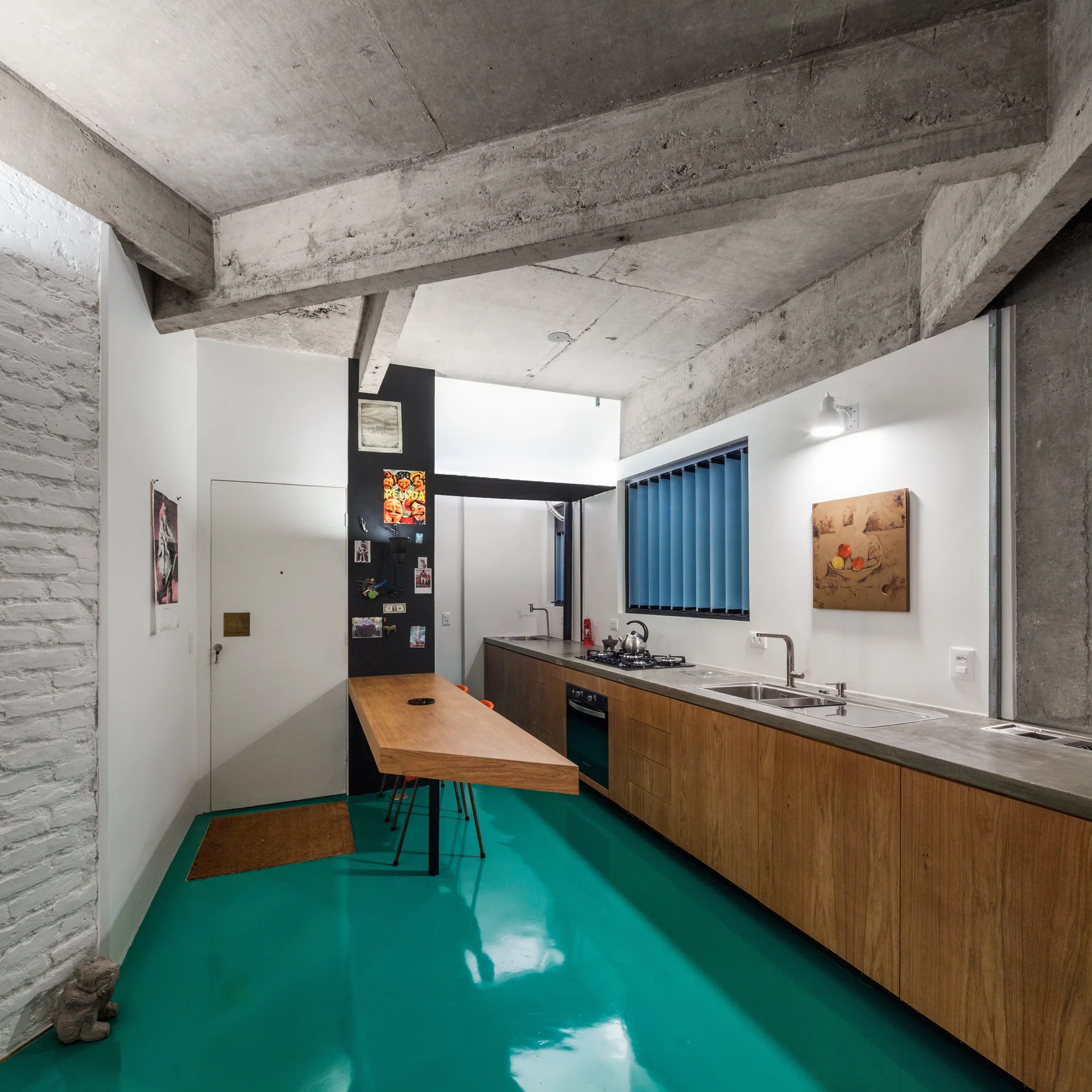 An industrial kitchen features a glossy teal floor, wood cabinetry under a concrete countertop, and an exposed concrete ceiling with beams.