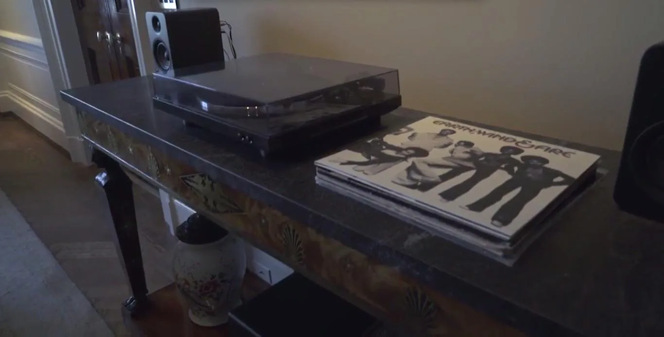 A dark grey stone-topped console table with carved wood detailing holds a record player, a black speaker, and a stack of vinyl records.