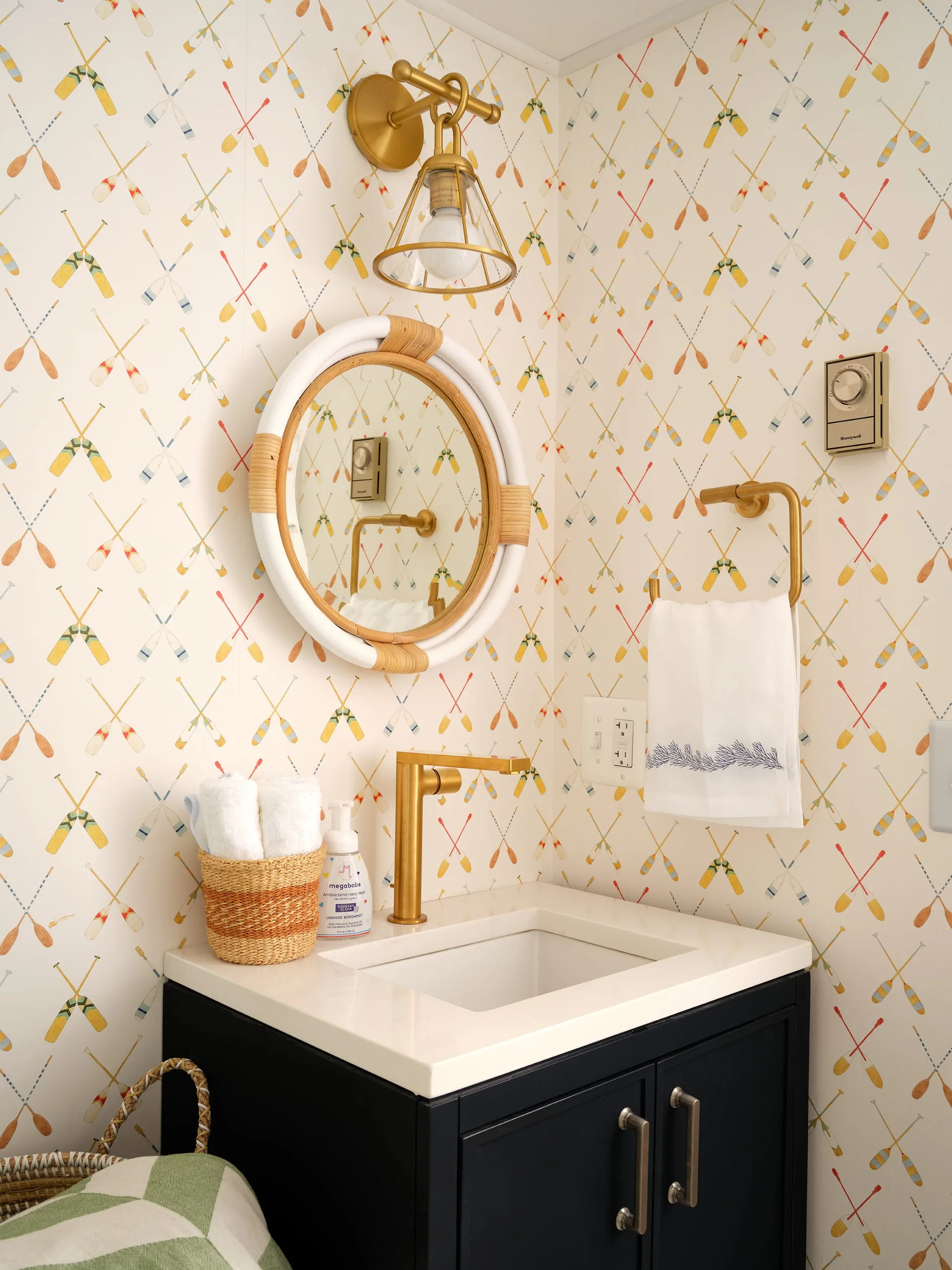 A dark blue vanity with a white stone countertop, a reed-wrapped round mirror, and brass fixtures against oar-patterned wallpaper.
