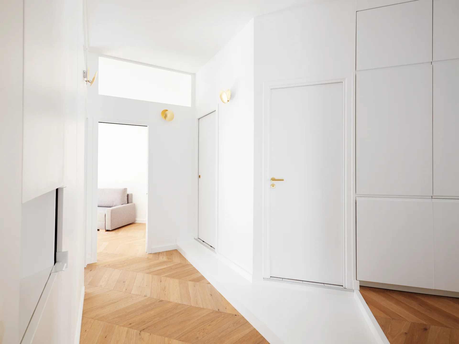 A bright white minimalist hallway features chevron patterned light wood flooring, flush white doors with gold handles, and round gold wall sconces.