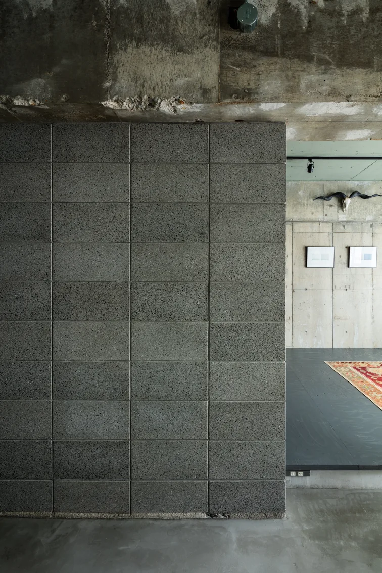 An interior view of a brutalist space featuring a dark grey concrete block partition wall, raw concrete floors, and distant framed artwork.