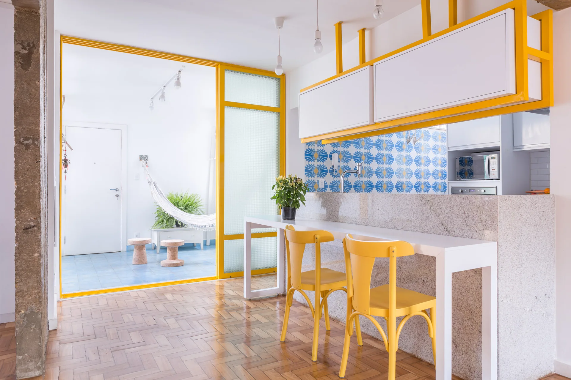 A modern kitchen features yellow chairs at a breakfast bar, white cabinets in a yellow frame, a blue geometric backsplash, and a sliding glass partition.