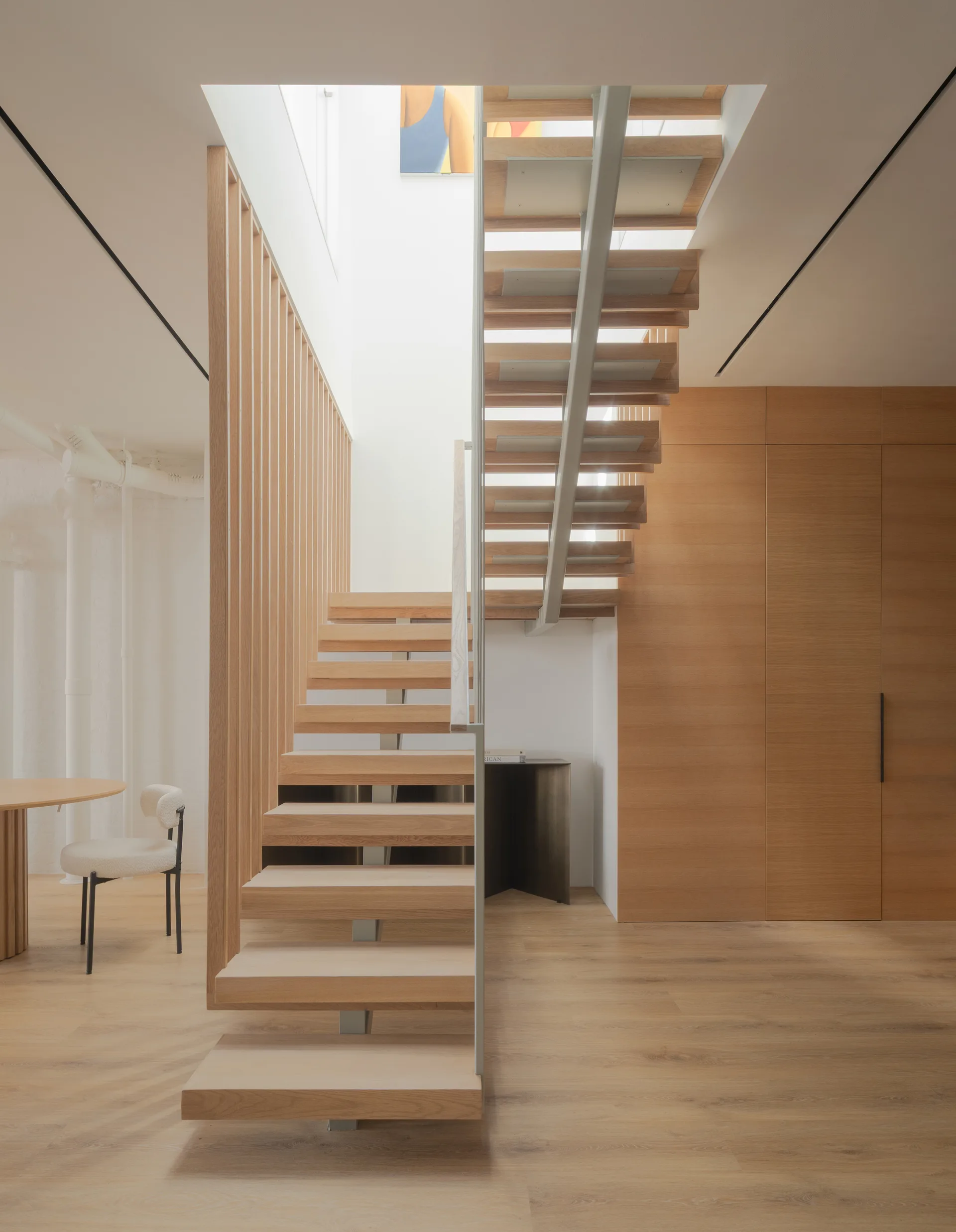 A minimalist floating wood staircase features open risers, a vertical slat screen, and flat-front wood wall paneling beneath a bright skylight.