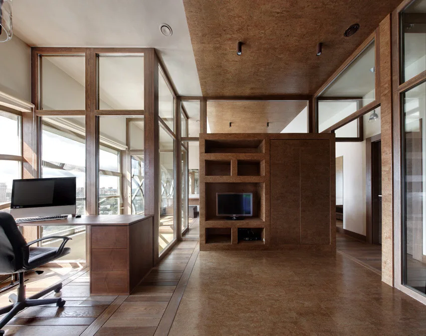 A modern home office featuring a wooden corner desk, a black rolling chair, and a central storage unit surrounded by glass partition walls.