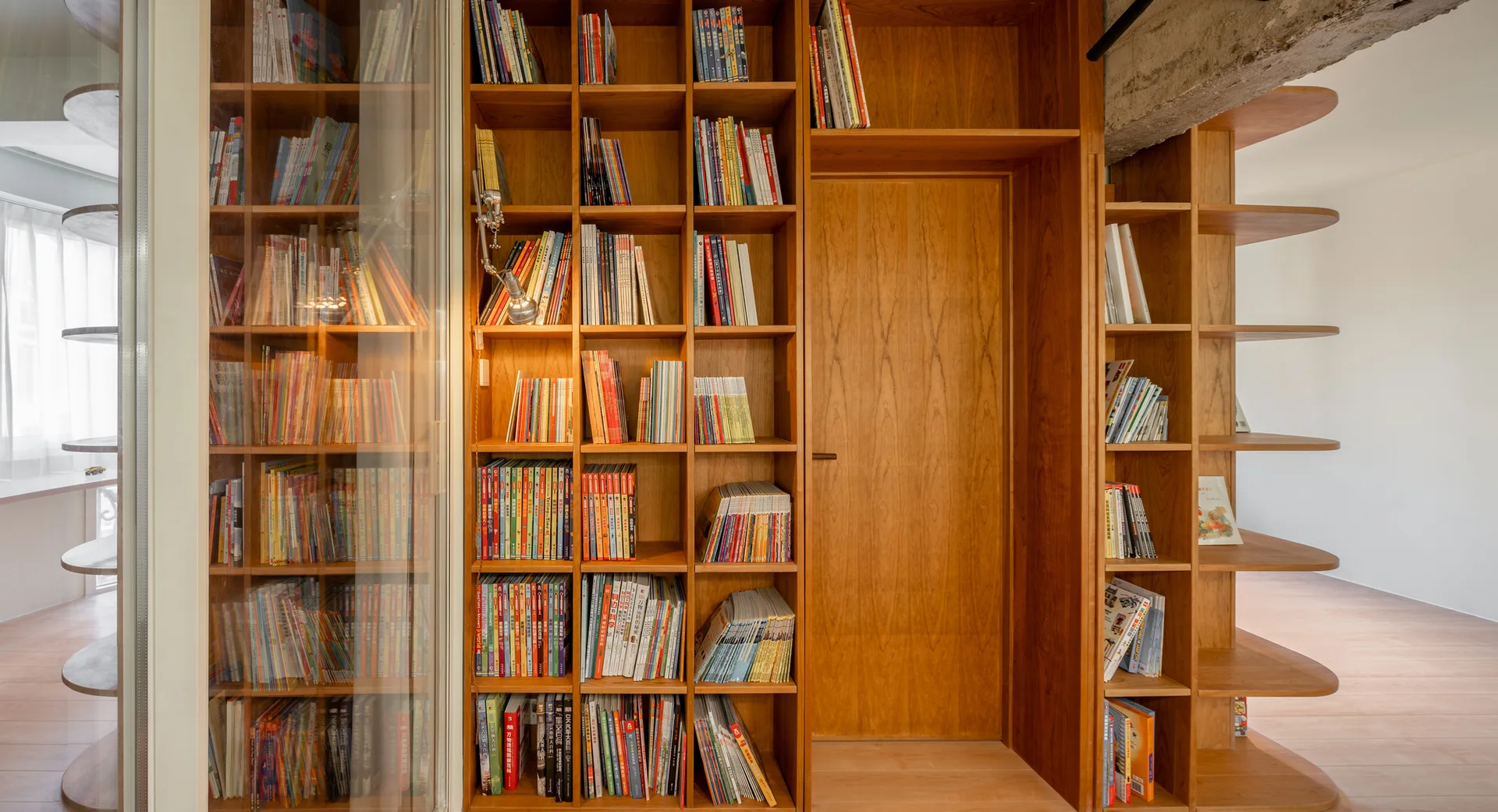 A large wooden bookcase wall with books features a flush integrated door and rounded end shelving under an exposed concrete beam.