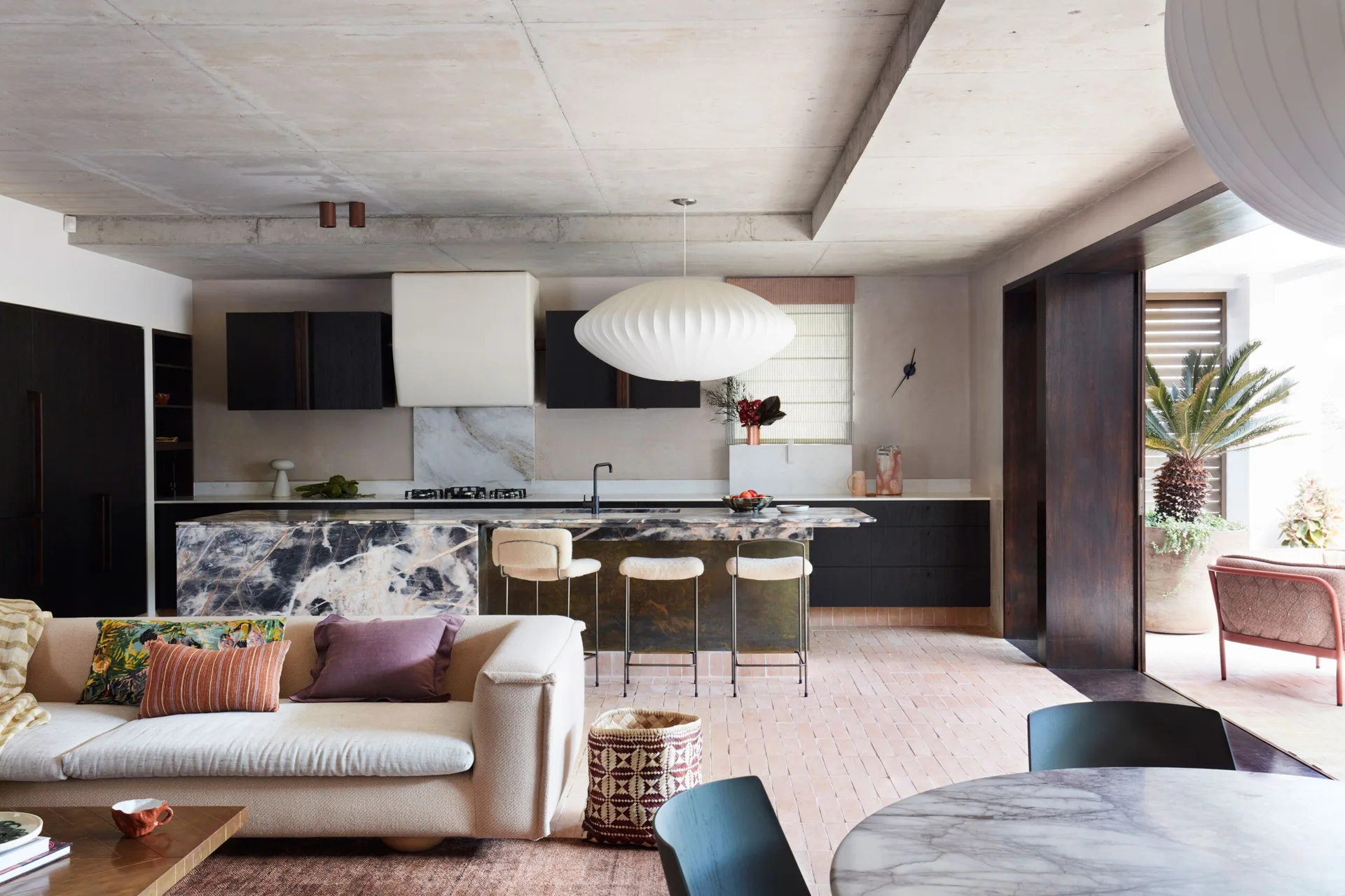 An open-plan kitchen and living space featuring a heavily veined marble island, dark timber cabinetry, and an exposed raw concrete ceiling.