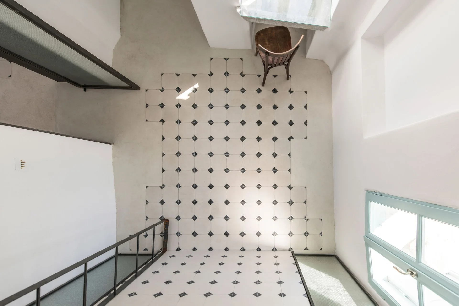 An overhead view of a bright stair landing featuring black and white patterned floor tiles, a dark wooden chair, and a metal stair railing.