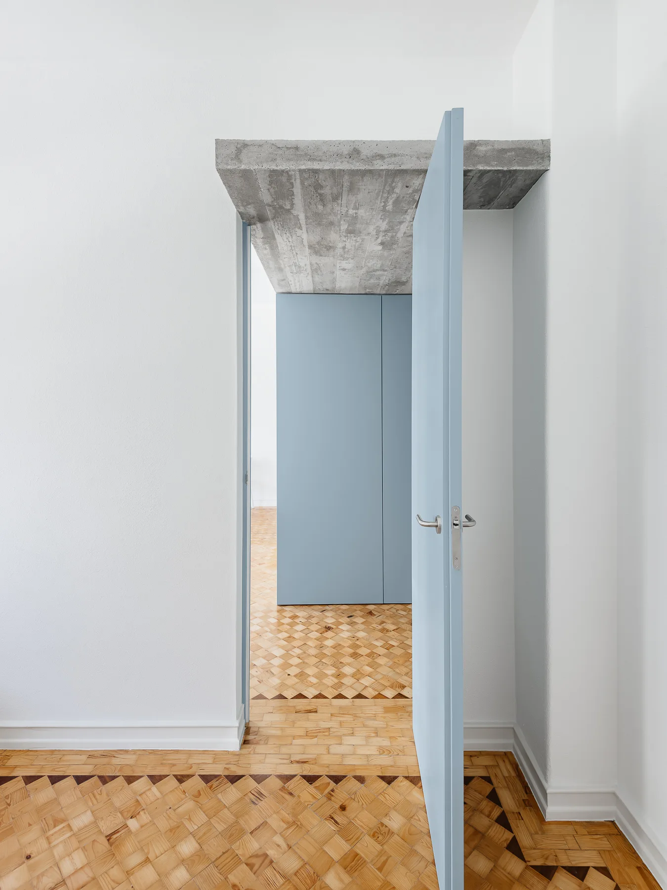 An open light blue door stands beneath an exposed concrete lintel in a room with geometric parquet wood floors and white walls.
